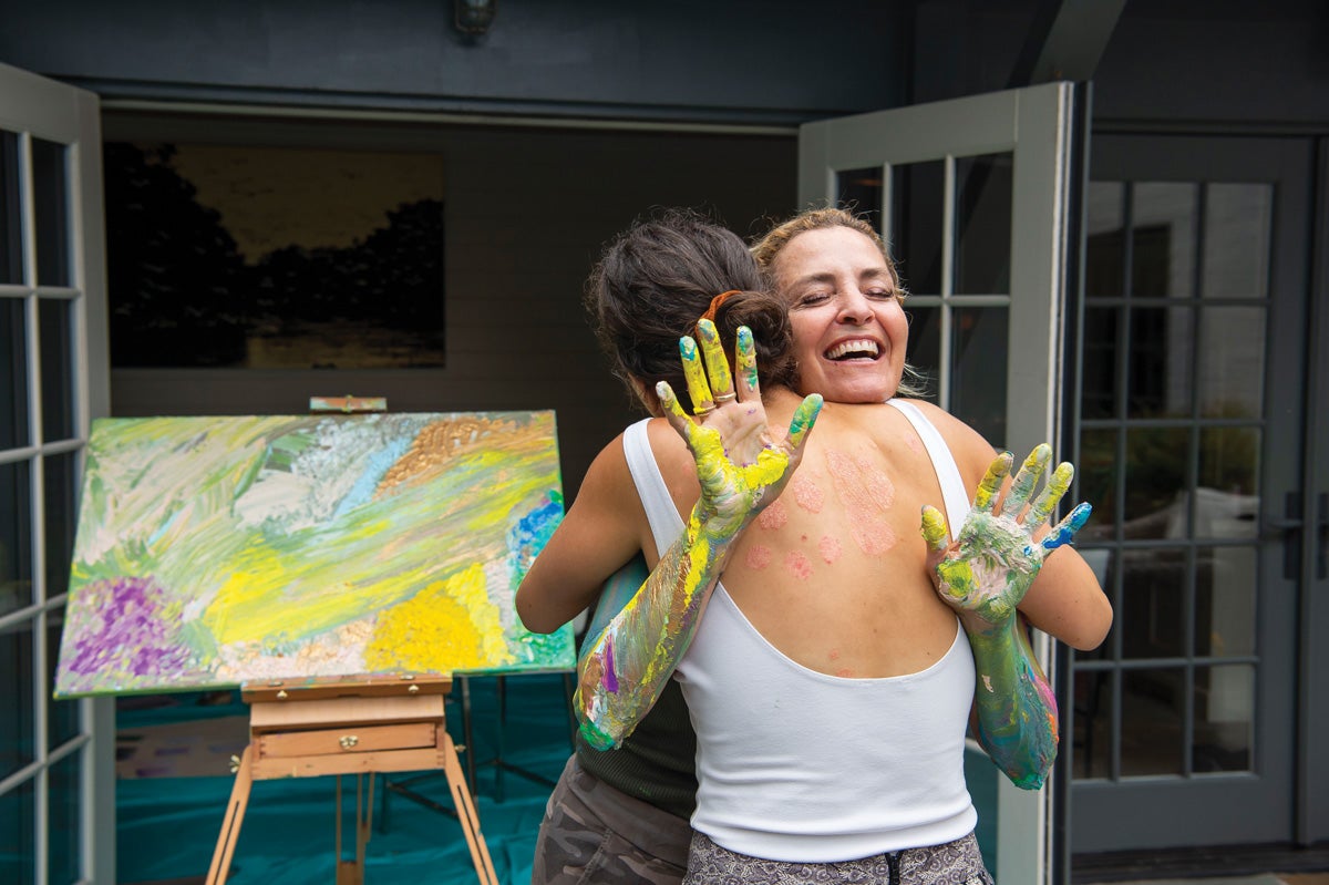 Two women hug each other in front of a painted easel. One of the women facing the camera holds out her painted hands.