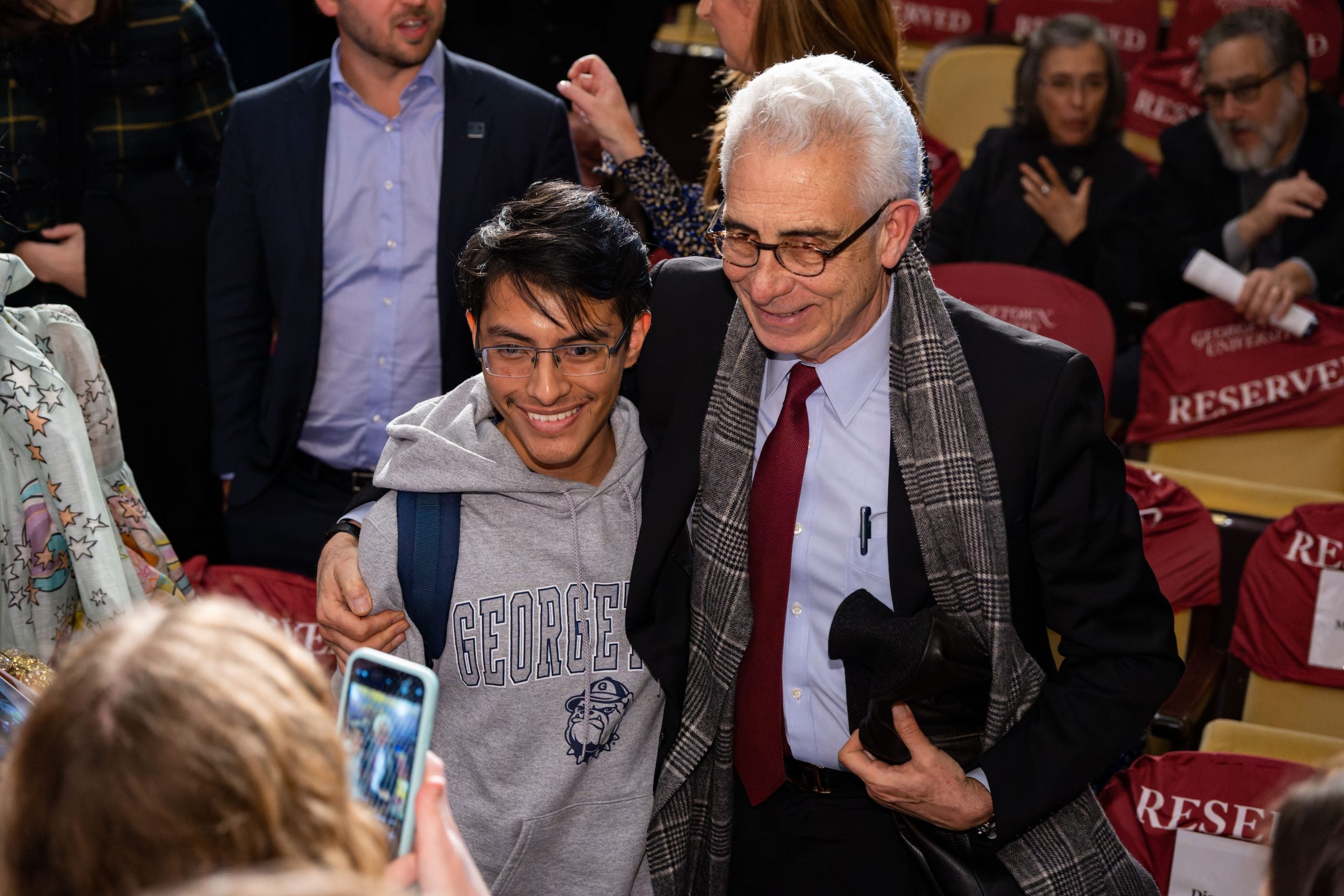 A former Latin American president (right) dressed in a black suit and red tie poses for a picture with a Georgetown student (left) wearing a gray sweatshirt that says "Georgetown."