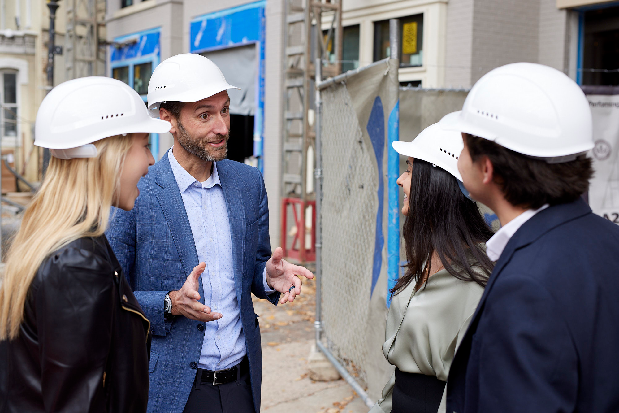 Two men and two women stand in a circle talking. They are dressed in formal business wear and all are wearing white hard hats. Behind them is a construction zone.