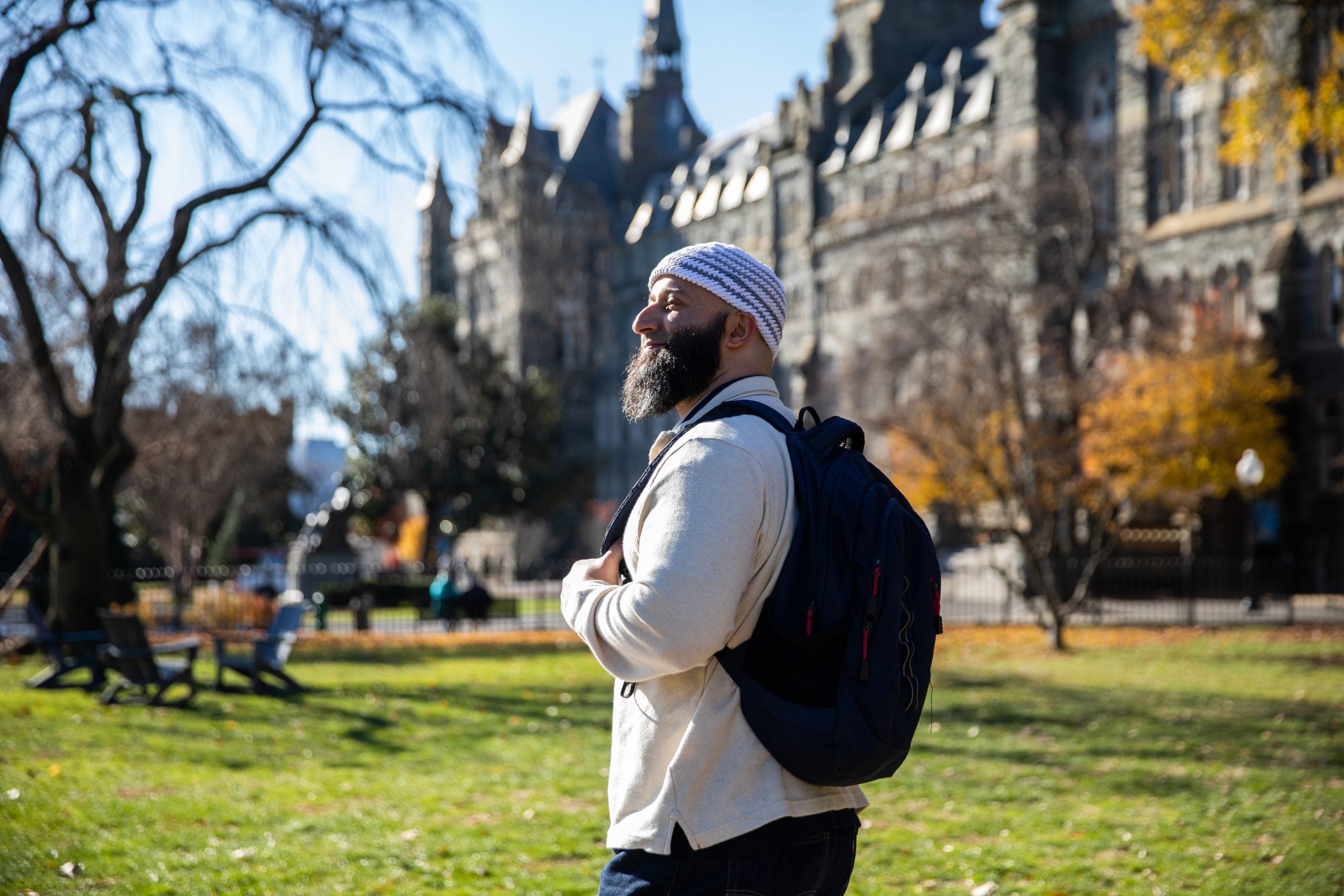 Adnan Syed, a program associate in the Prisons and Justice Initiative, wears a cream-colored sweater, a blue-and-white-striped kufi, or Muslim prayer hat, and a black backpack. He stands on the front lawn of Georgetown with Healy Hall, a Gothic-style building behind him and looks forward.