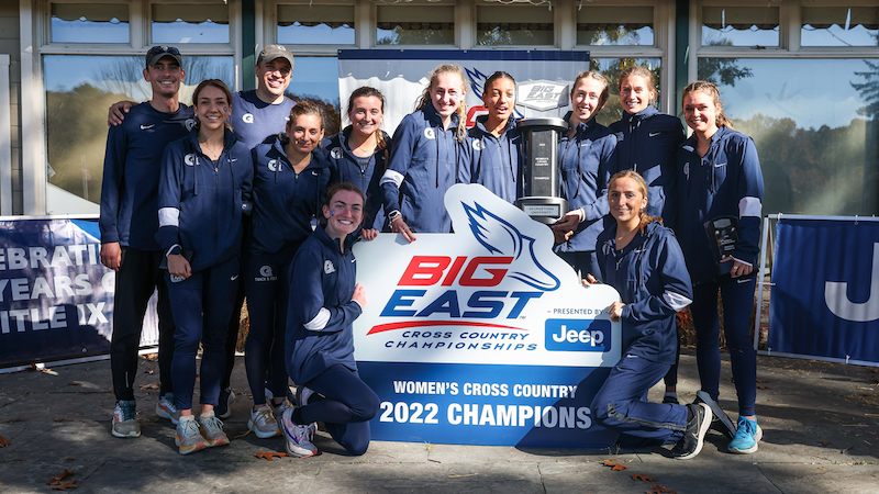 Women's cross country team in dark blue sweat suits stand behind a Big East 2022 Champions sign