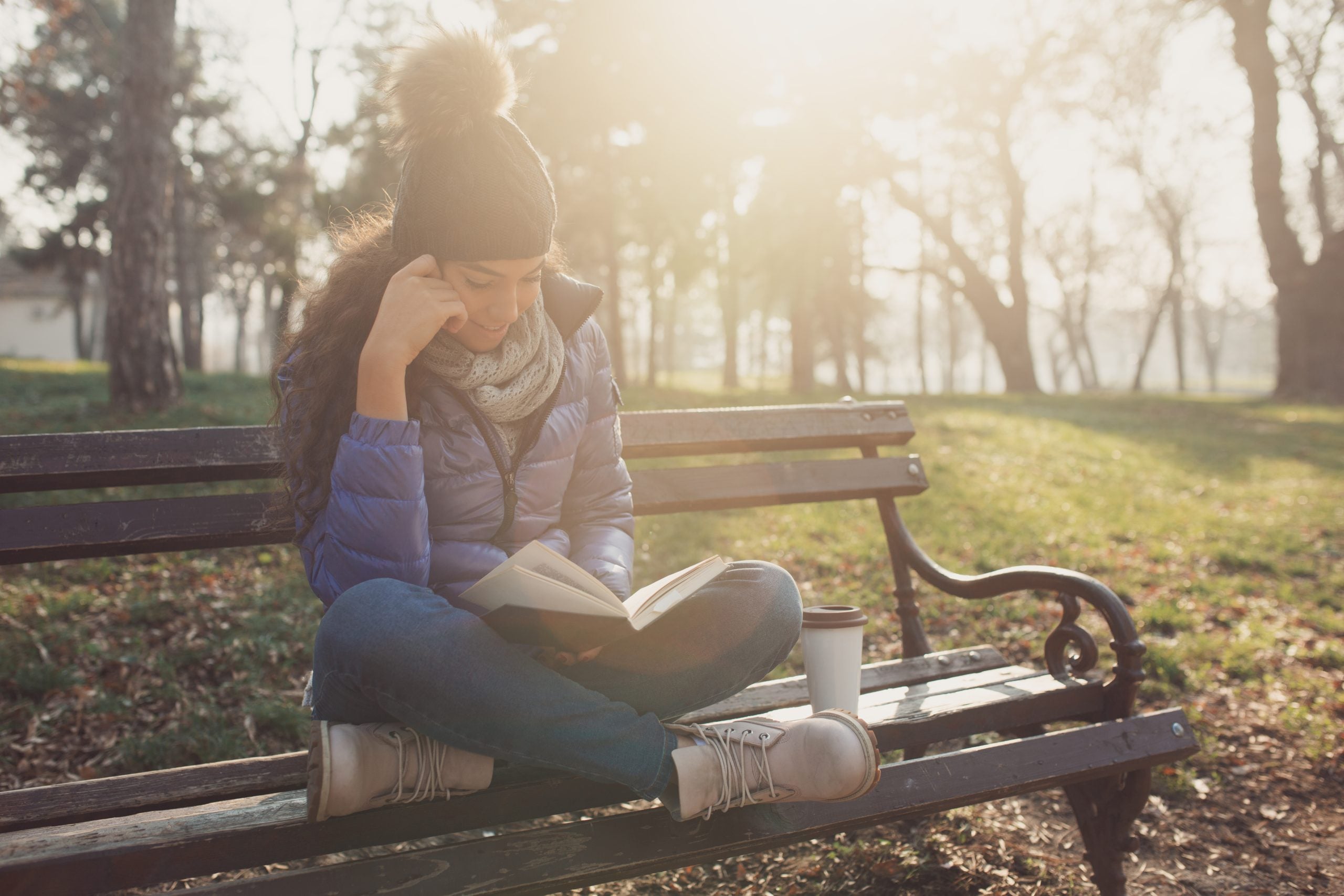 A young woman sits on a park bench cross-legged reading a book. She is wearing a pompom hat, a purple winter jacket and jeans. Next to her on the bench is a coffee cup.