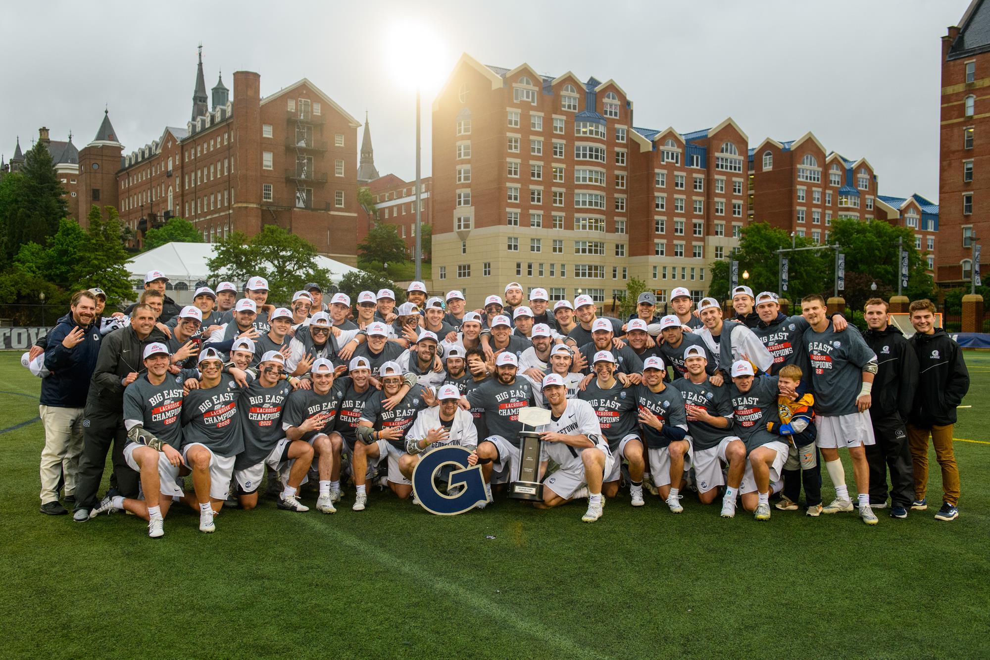 Men's Lacrosse team celebrates in a group photo with the sun setting behind the field