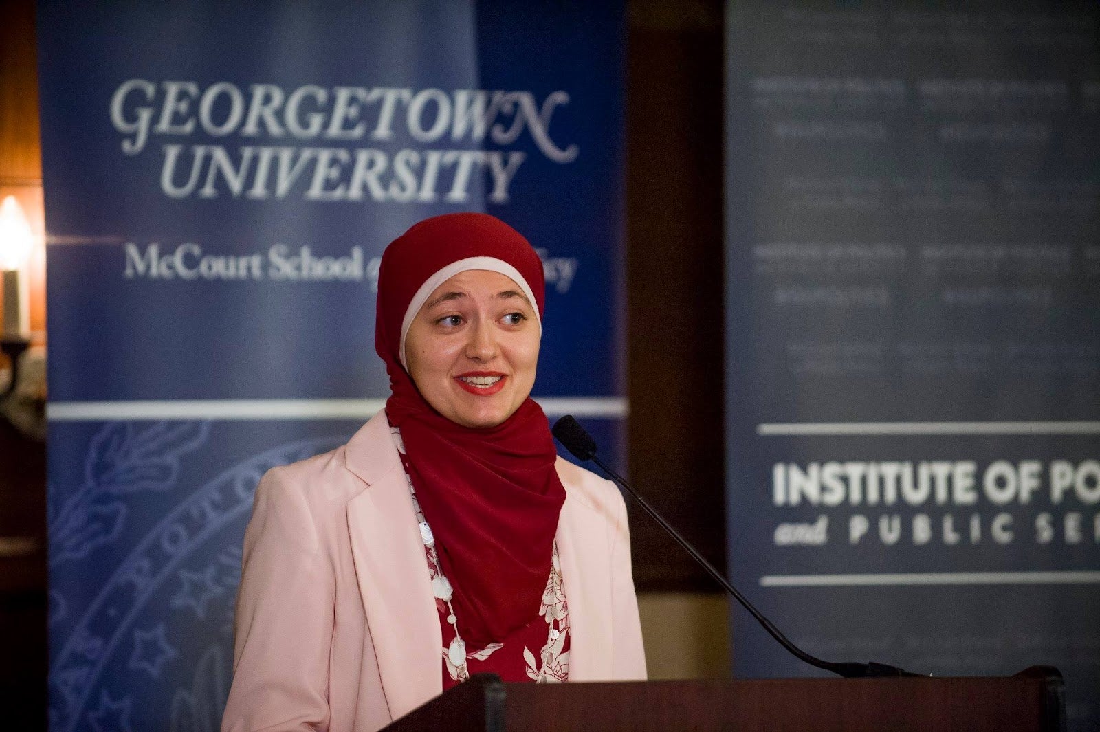 Ruwa Romman, a graduate of the McCourt School, wears a red hijab and a pink suit while speaking at a podium in front of a blue sign that says "Georgetown University."