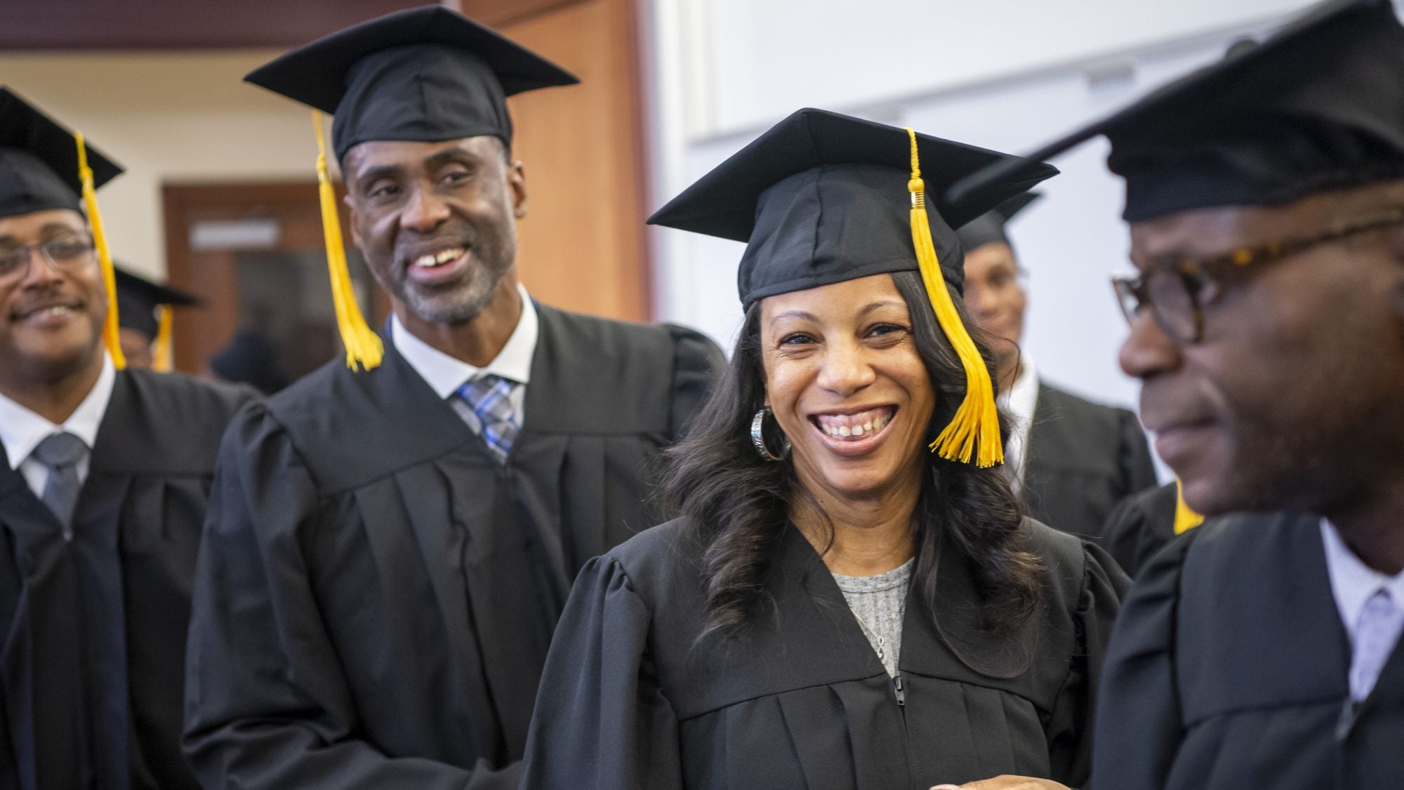 A group of returning citizens wear black graduation robes and caps with yellow tassels dangling on one side. They are smiling during an event celebrating their graduation from Georgetown's Paralegal Program.