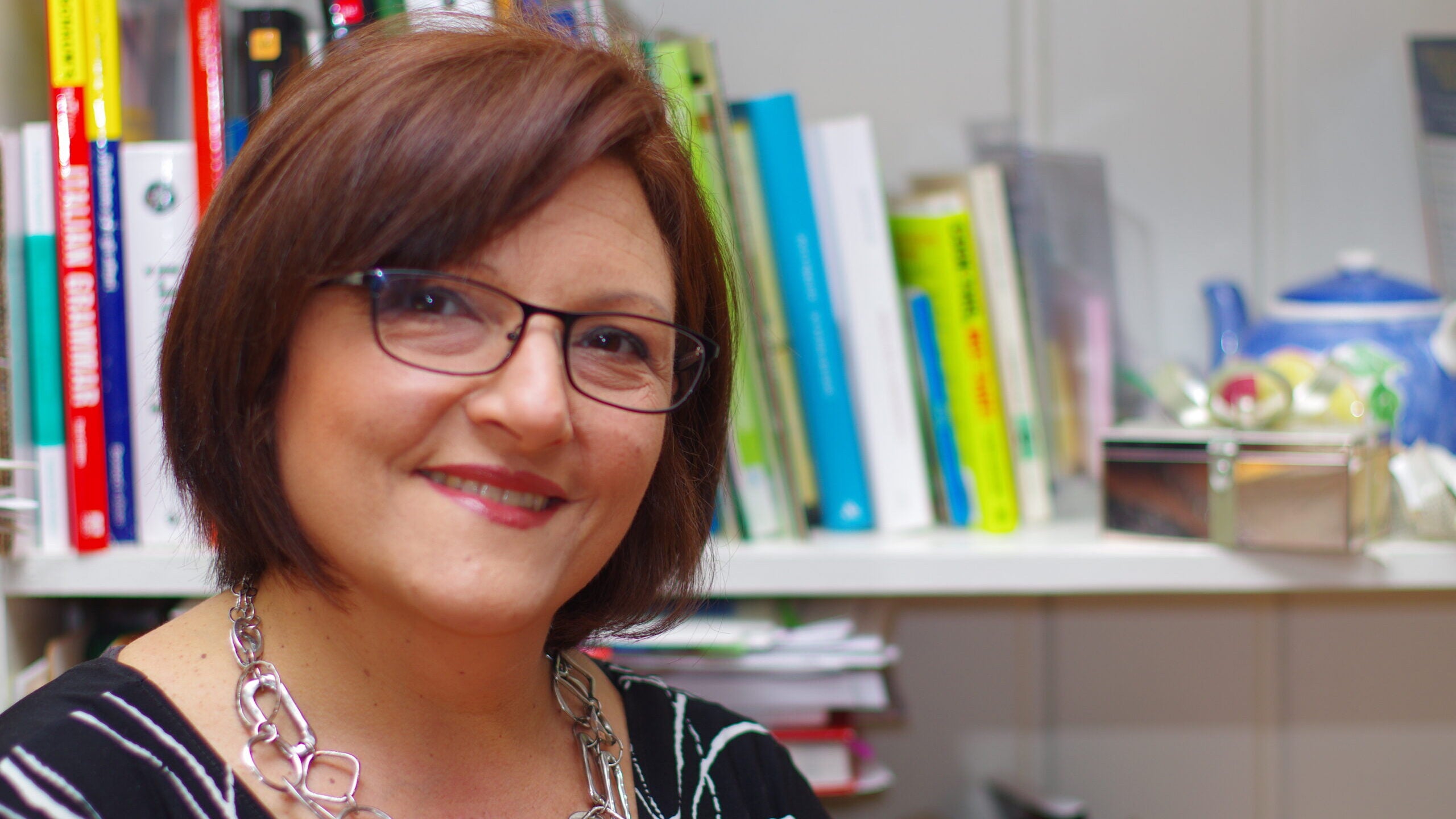 Donatella Melucci, an Italian language professor, is pictured sitting in front of a bookshelf. She has short auburn hair, rectangular glasses, a black shirt and a chunky silver necklace.