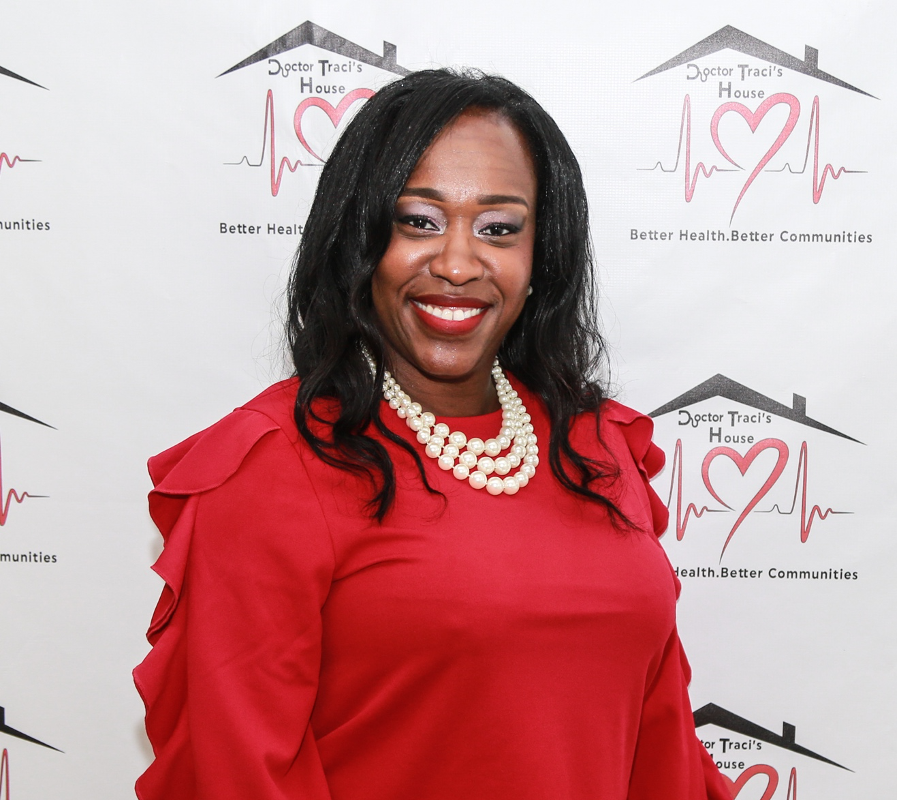 Dr. Traci Thompson (C'96) stands in front of a white background with her clinic's logo "Dr. Traci's House" written behind her. She has long black hair and wears pearls, a red long-sleeve shirt, and a black skirt. Her nails are painted pink.