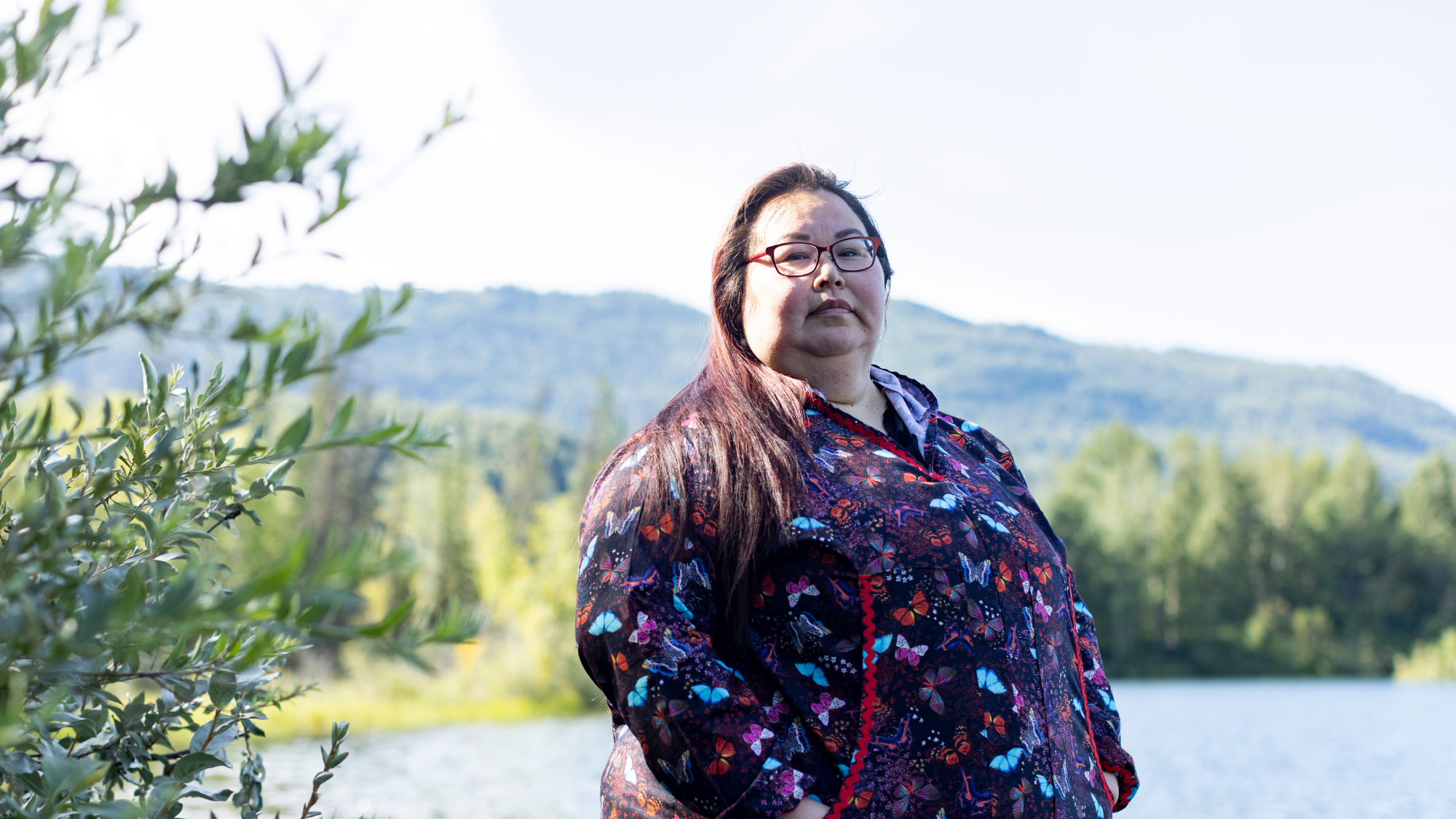 Angel Dotomain (B'99) wears a purple button-up shirt with blue, red, and multi-colored butterflies on it and rectangle glasses. She stands in front of a green mountain, which is blurred behind her.