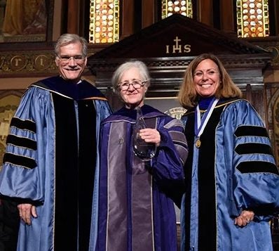 Professor Maxine Weinstein (center) wears a purple graduation robe at a commencement. She stands on the stage of Gaston Hall next to Provost Robert Groves, who wears a blue graduation robe with three black stripes on the sleeves, and another woman wearing a blue graduation robe with three black stripes on the sleeves.