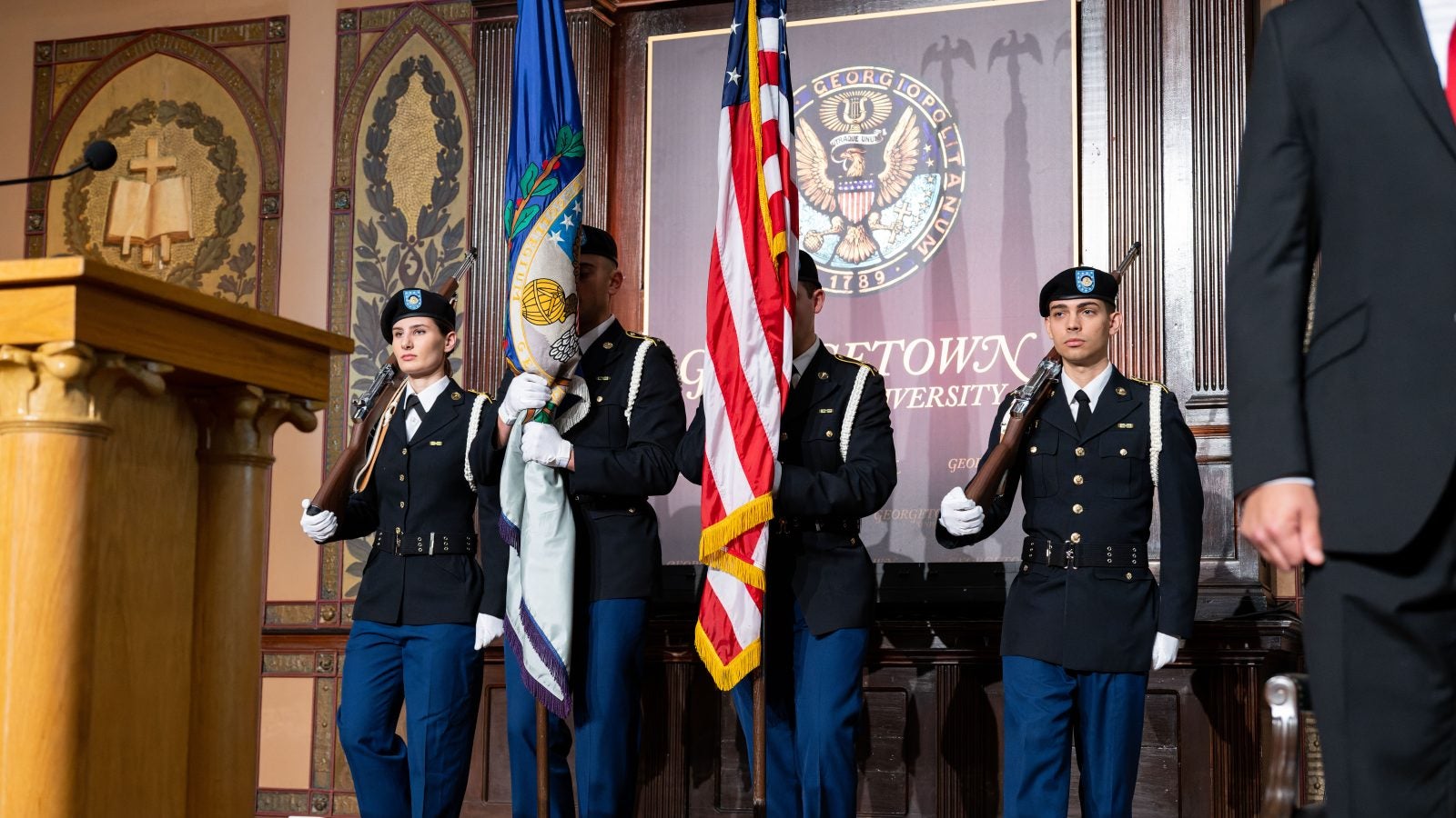 Students wearing military regalia stand on stage in Gaston Hall, holding an American flag.