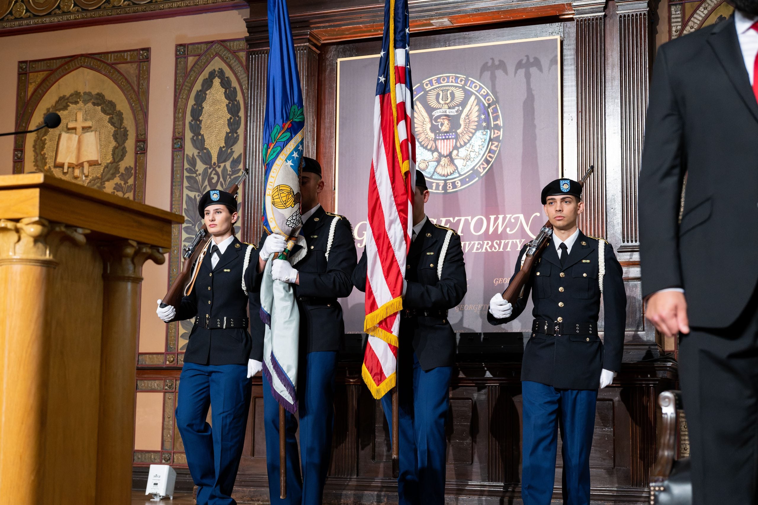 Students wearing military regalia stand on stage in Gaston Hall, holding an American flag.