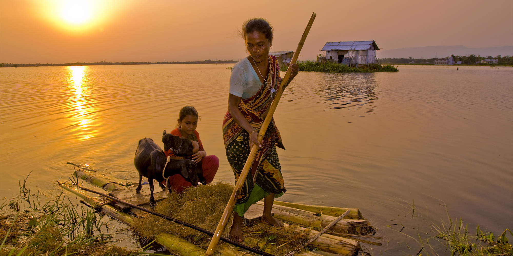 A woman, child, and goats stand on a boat constructed out of wood. The boat floats in a body of water at sunset as the mother uses a wooden stick to maneuver.