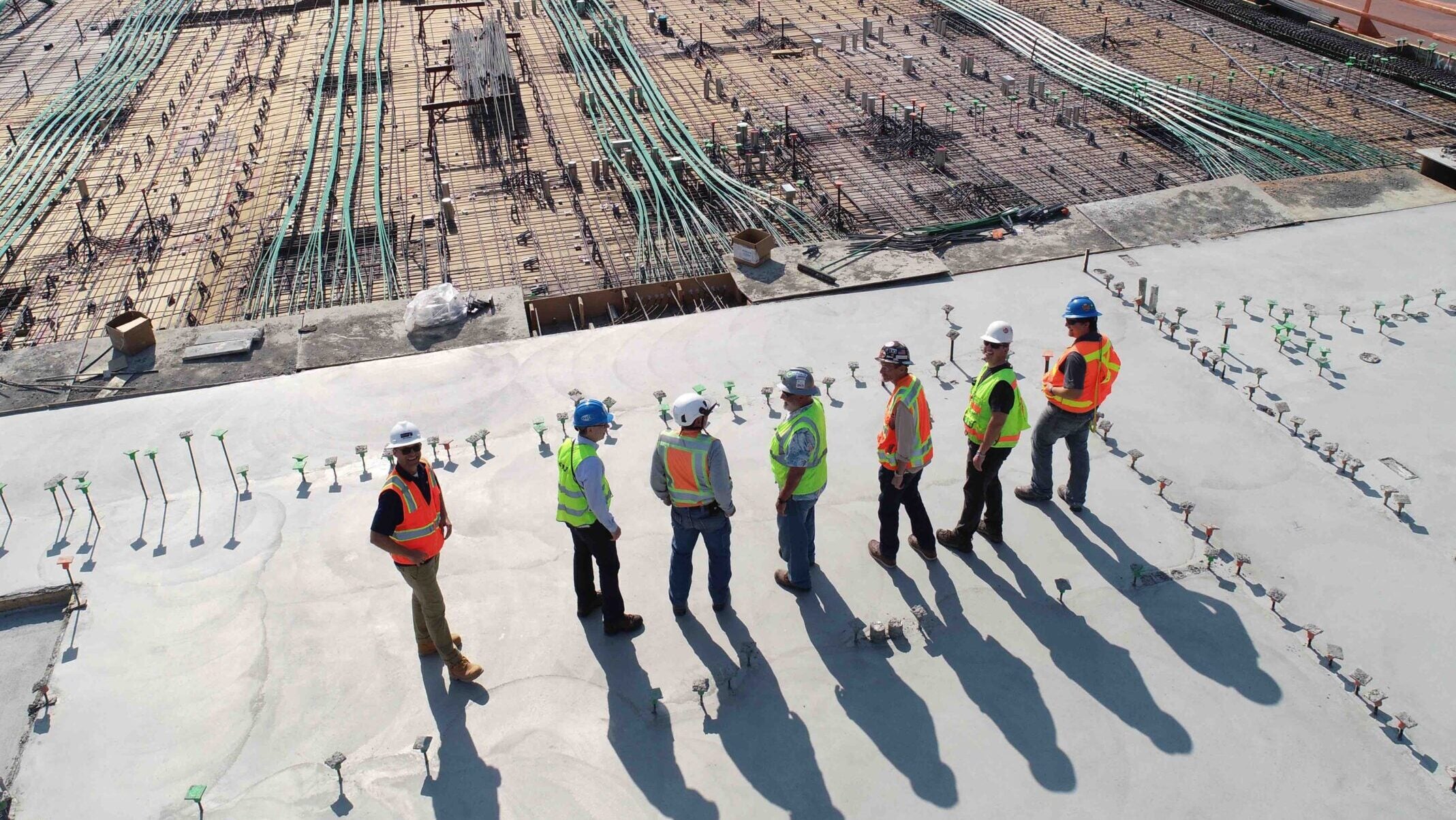 A view from above as seven construction workers stand on a cement block on a sunny day.