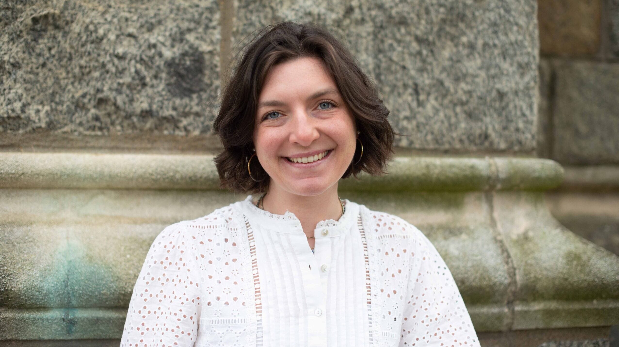 Grace Buono, a senior who just finished her thesis on the metaverse, stands in front of a stone wall in a white shirt and smiles.