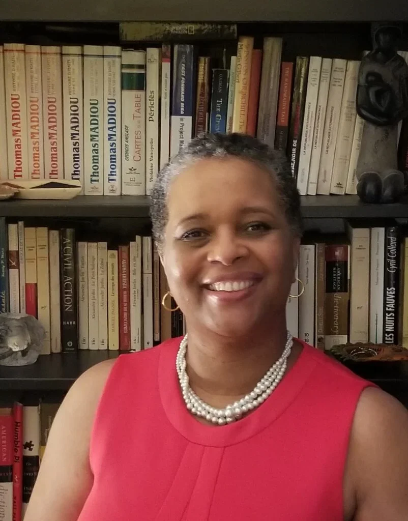 Carline Noailles (EMBA'23) smiles at the camera. She wears a bright pink sleeveless shirt and pearls and stands in front of a bookshelf.