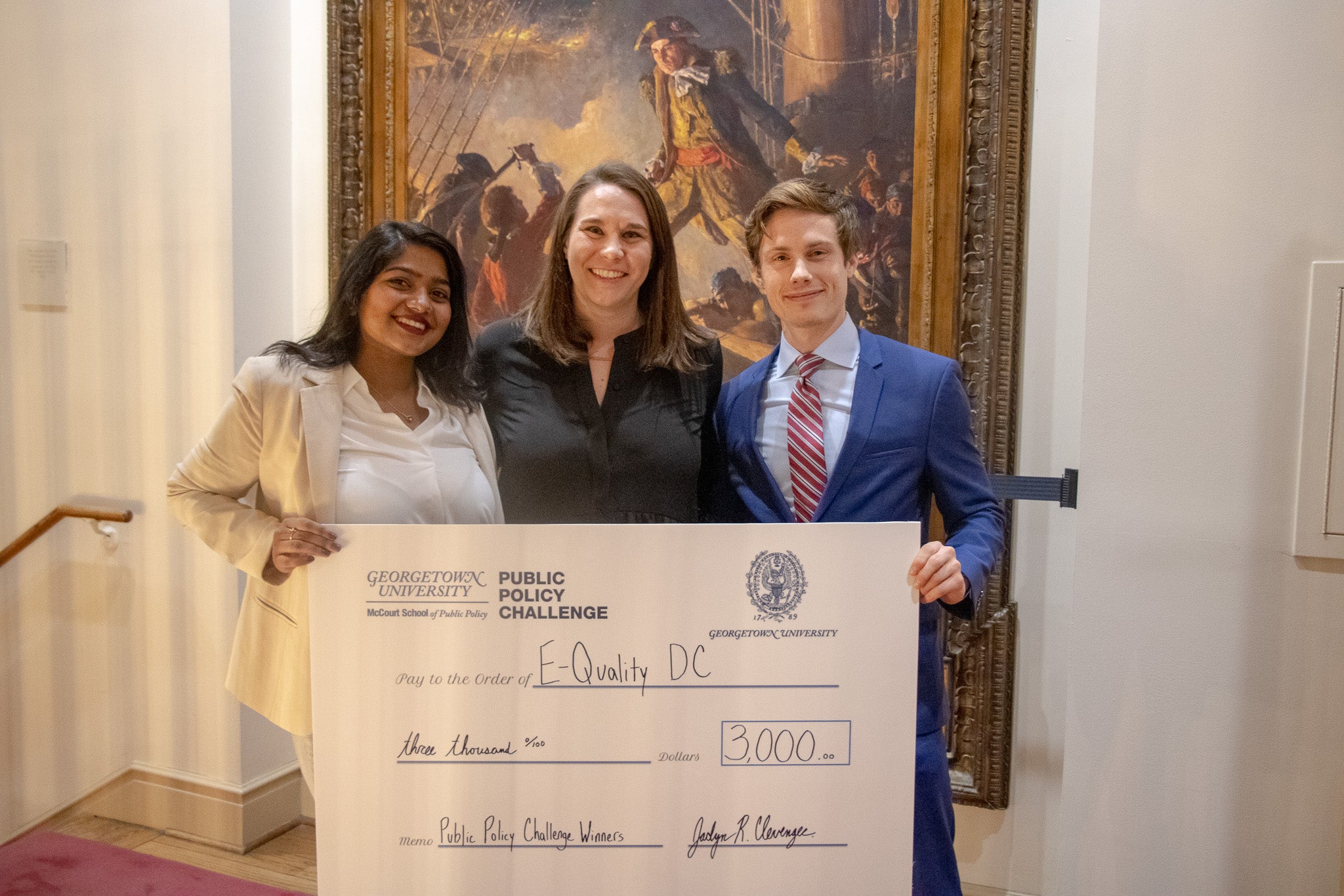 Three winners of the Georgetown Policy Challenge stand side-by-side holding a check. Behind them is an oil painting.