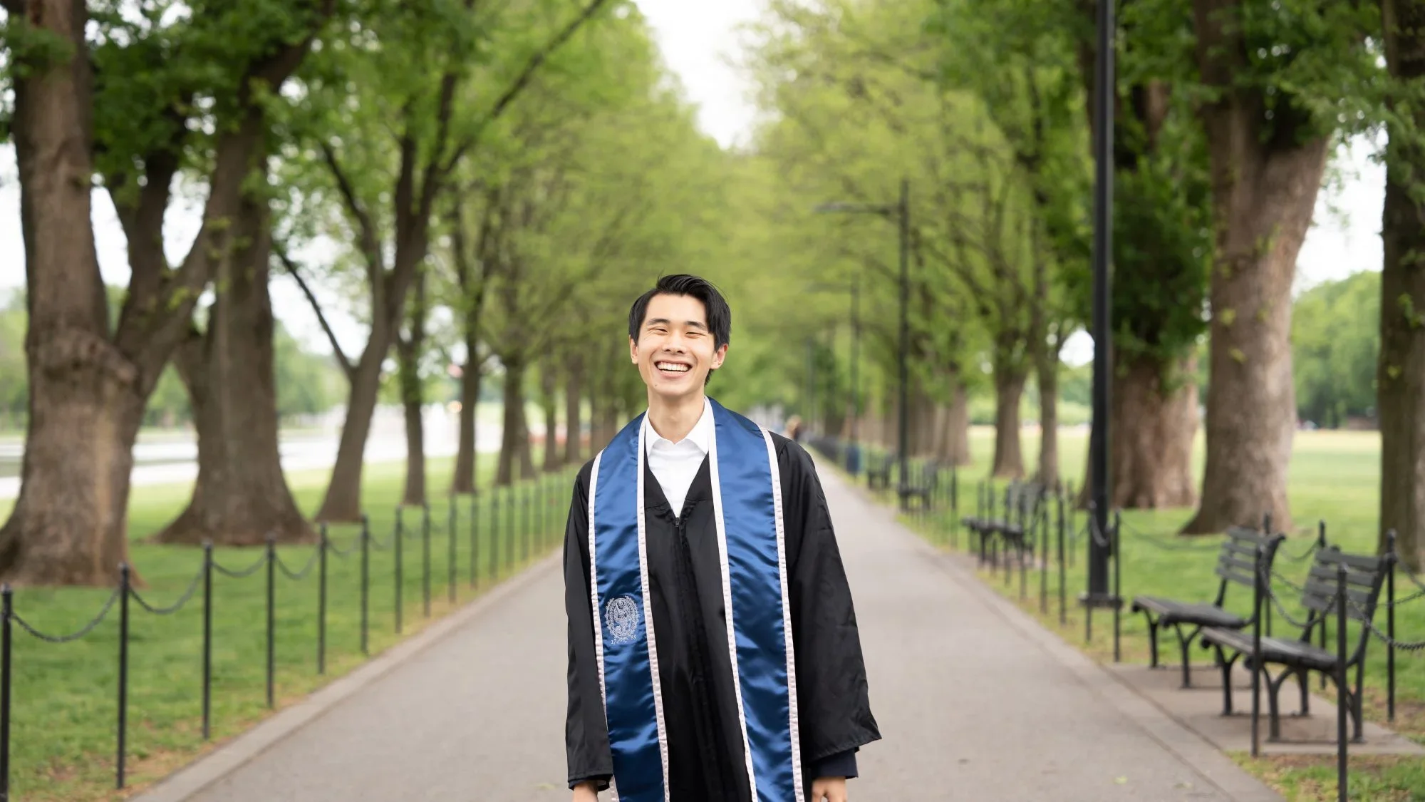 Max Zhang (SFS'23) poses in graduation gown and stole on a path canopied by trees.