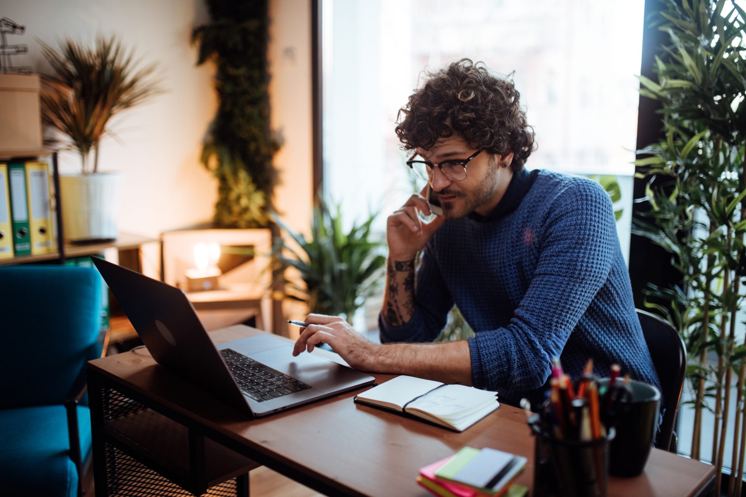 An employee talks on the phone while typing on his laptop at his home desk. Behind him are green plants.