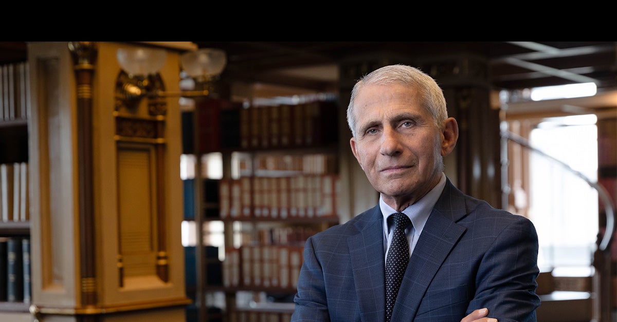 Dr. Anthony S. Fauci stands with his arms crossed in Riggs Library on Georgetown's Main Campus. He wears a navy suit with a white shirt underneath and tie, and stands in front of bookcases.