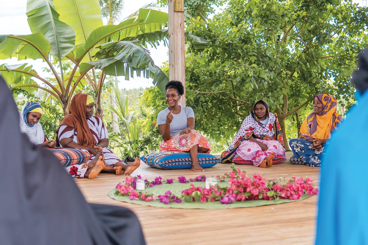 Certified nurse midwife Nafisa Jiddawi(NHS'14) sits on a pillow on the ground of a holistic wellness center dedicated to women's health promotion and disease prevention that she established in Tanzania. In front of her lays a circle of flowers; behind her are palm fronds.