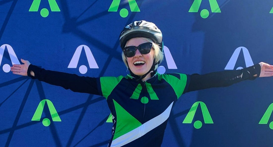 Marissa Nissley (B'24) stands at the finish line after a 50-mile race with her arms outstretched. She wears black sunglasses and a helmet.