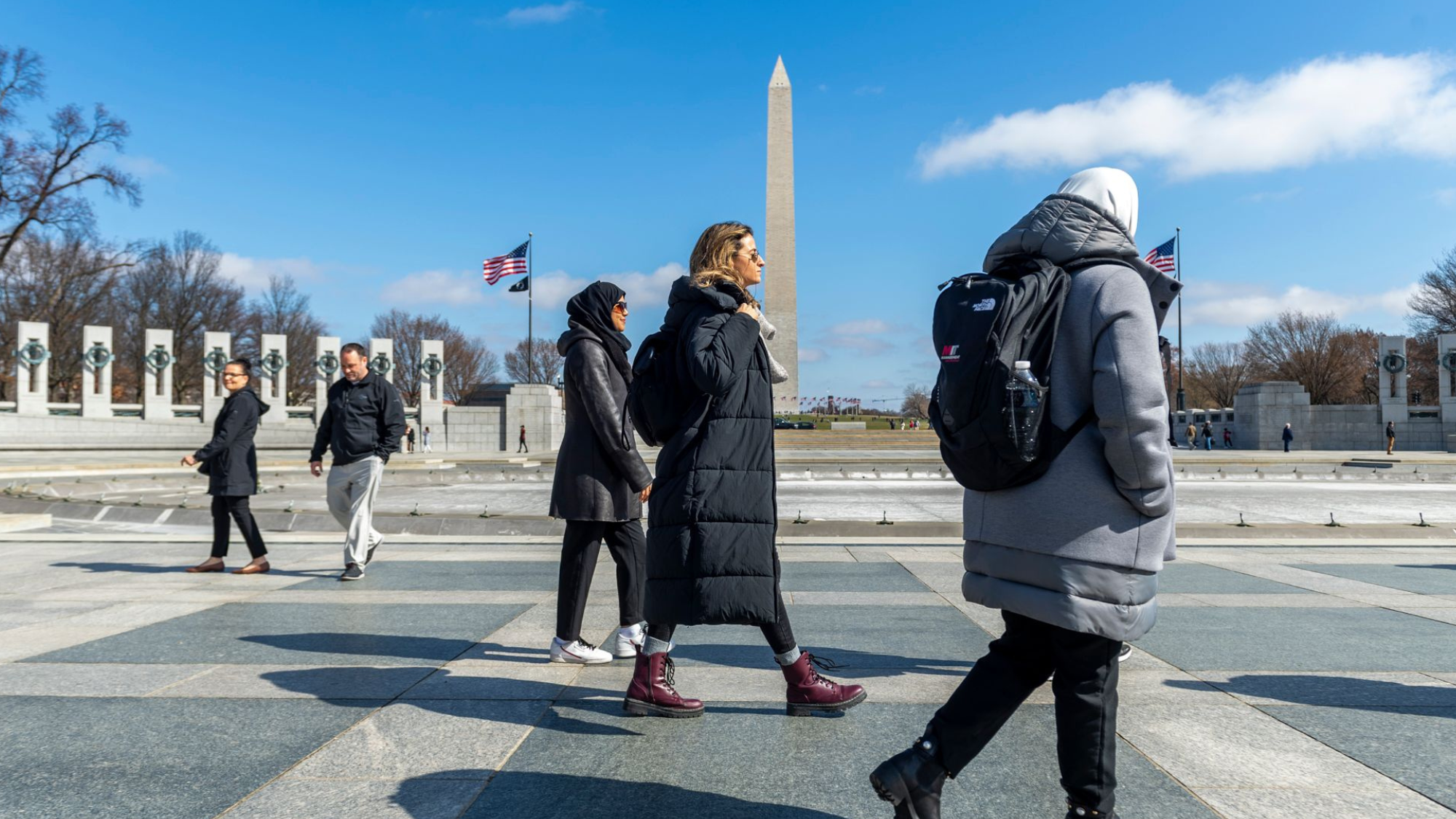 A group of people in jackets walk through the World War II Memorial with the Washington Monument in the background.