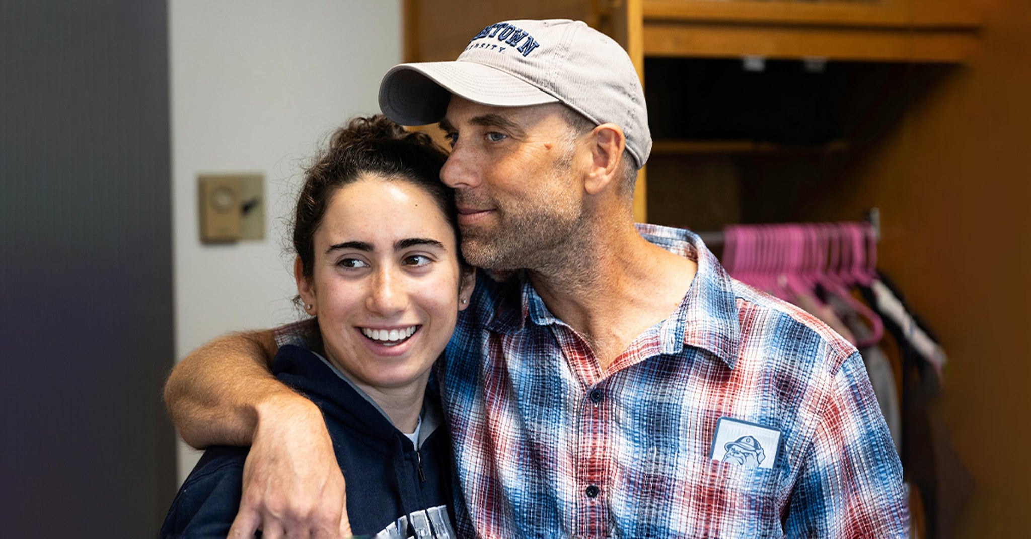 A father wraps his arm around his daughter and leans his head against hers. His daughter smiles off-camera.