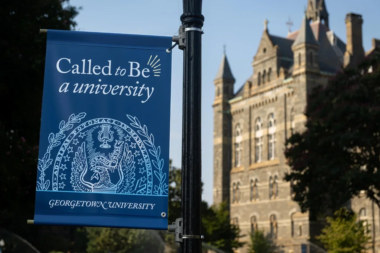 A blue flag hoisted on Georgetown's campus reads "Called to Be a university." Behind the flag is Healy Hall, a historic stone building on Georgetown's main campus.