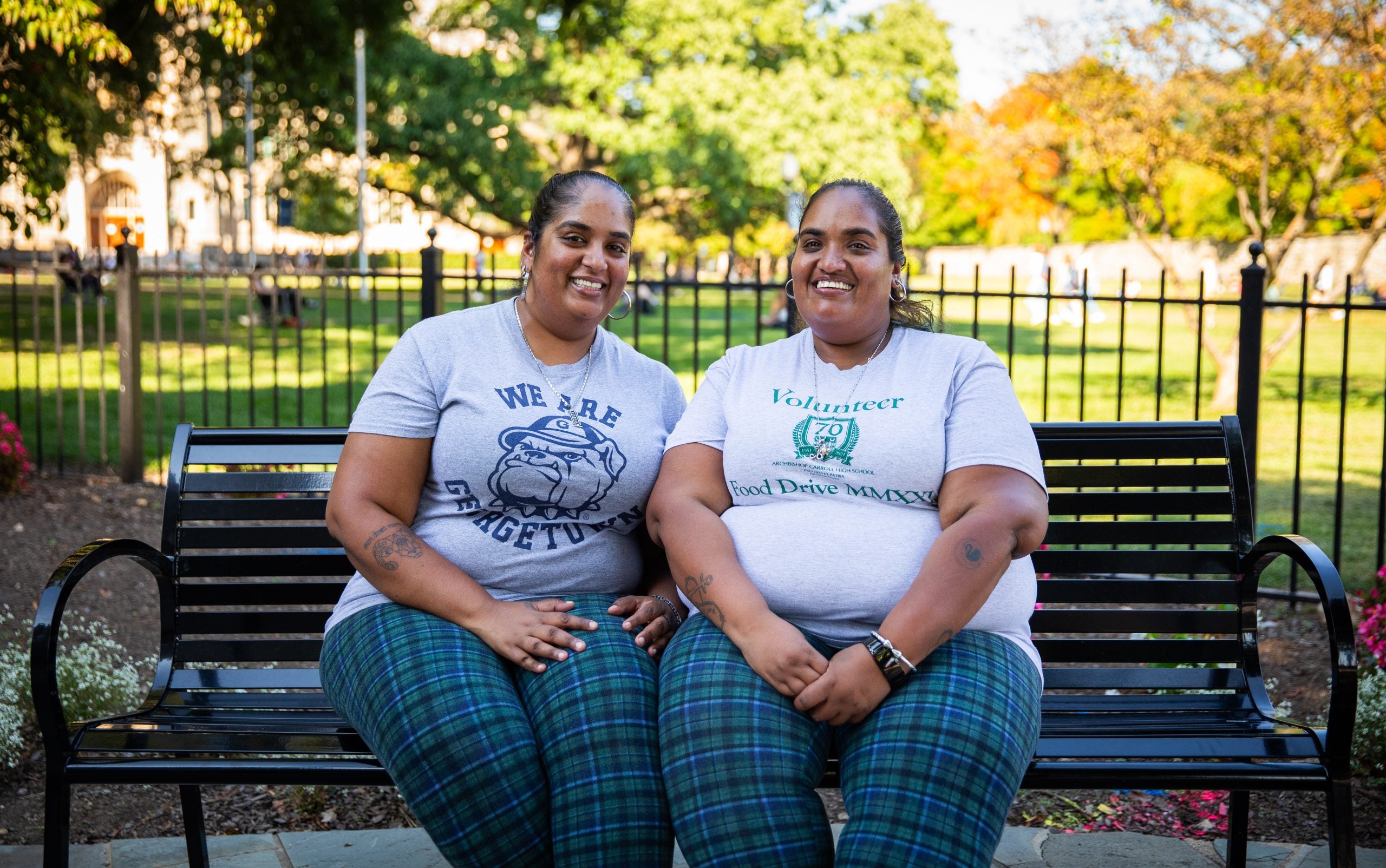 Two sisters wearing matching plaid pants and gray T-shirts sit on a park bench by the front lawn of Georgetown.