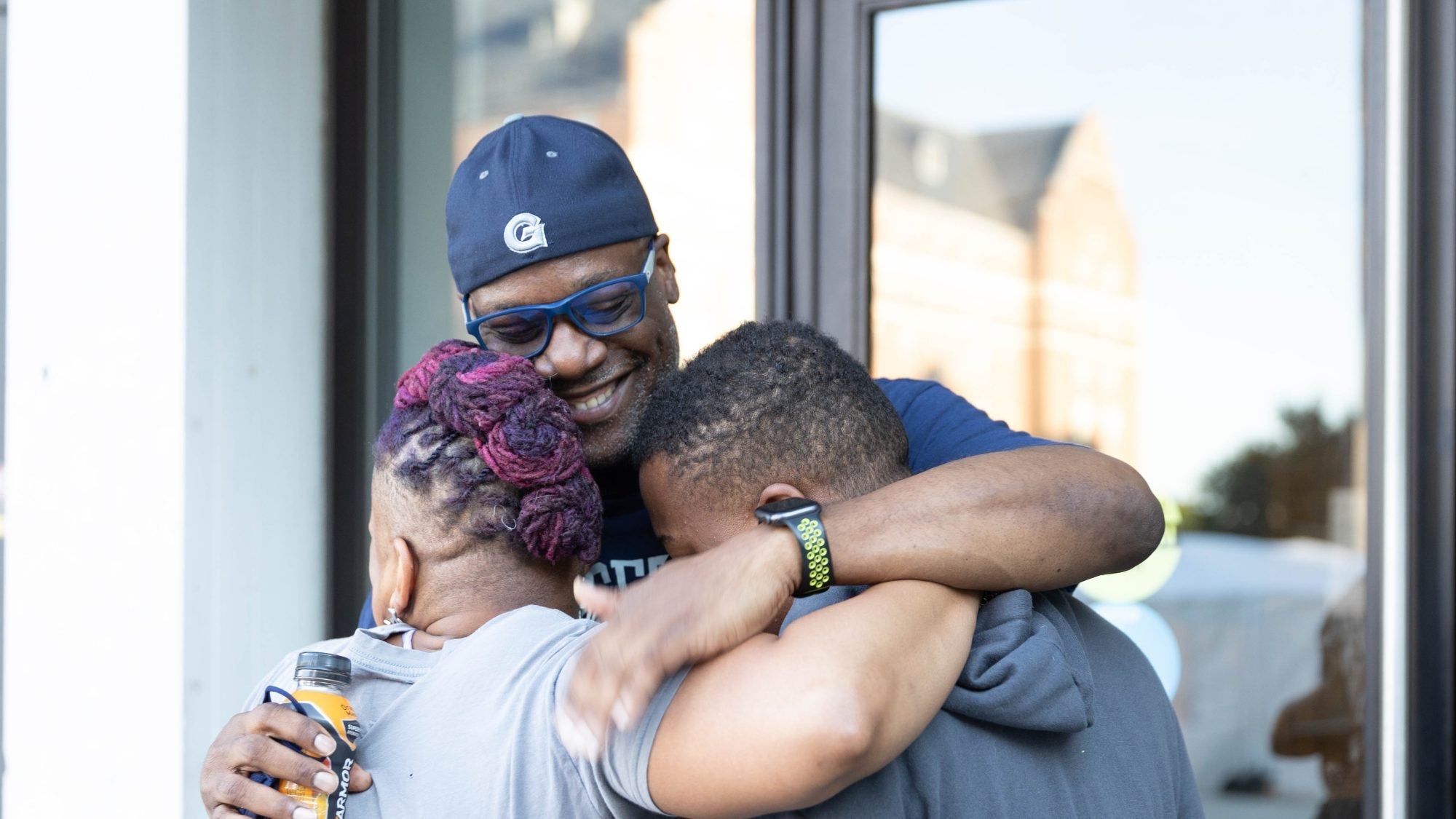 A parent hugs two children on Georgetown's campus wearing a Georgetown Hoyas baseball cap.