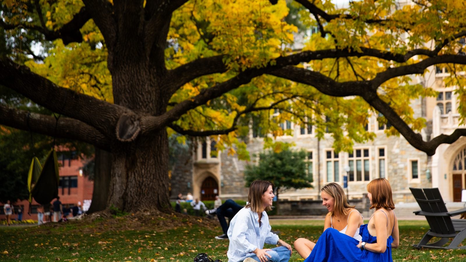 Three white female students sit on a blanket on the front lawn of Georgetown in front of a giant tree.