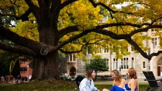 Three white female students sit on a blanket on the front lawn of Georgetown in front of a giant tree.