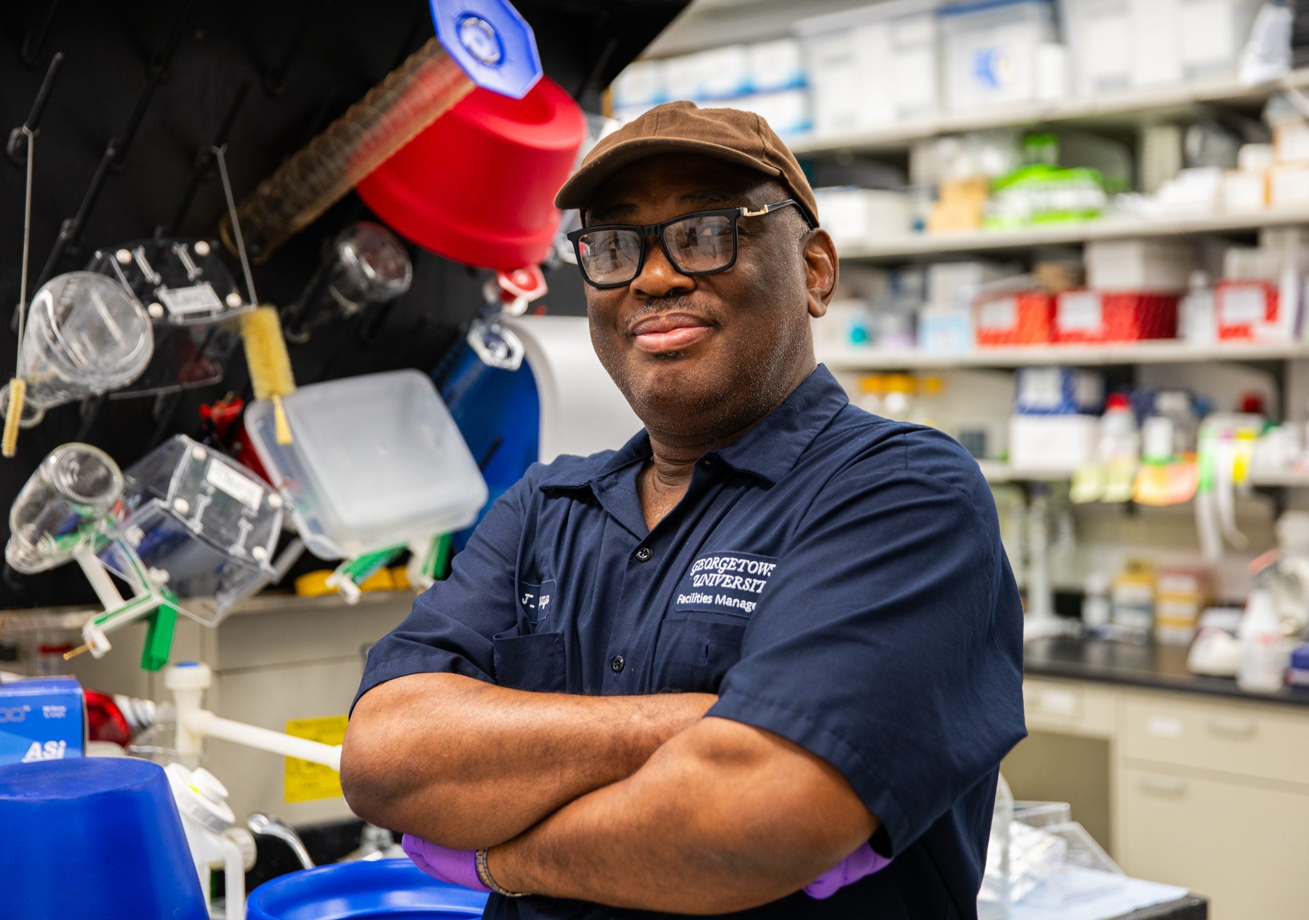 A custodian poses with his arms crossed and smiles slightly at the camera. He wears a navy uniform, a tan baseball cap and black glasses.