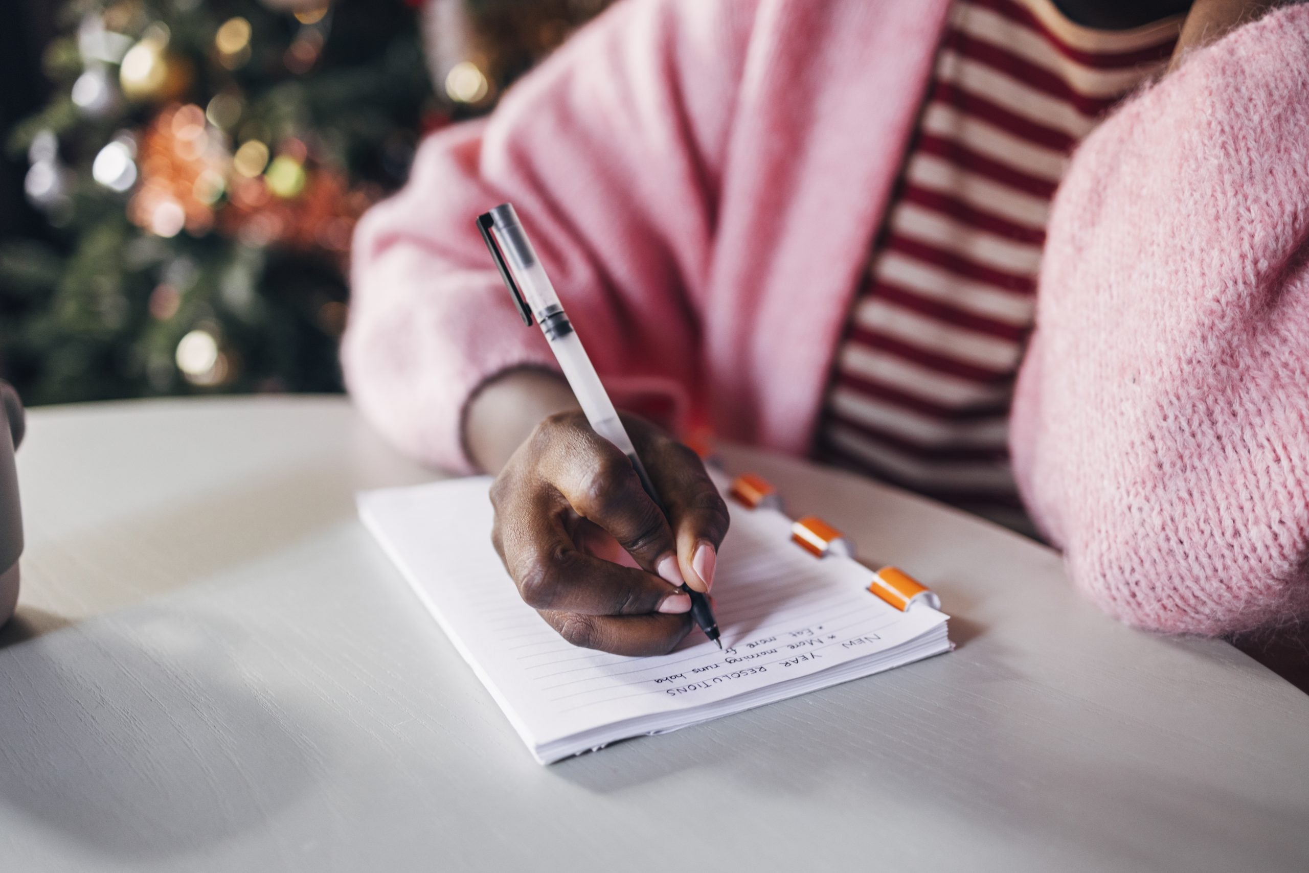 A Black woman wearing a pink sweater and striped pink shirt writes her New Year's resolutions on a notebook.