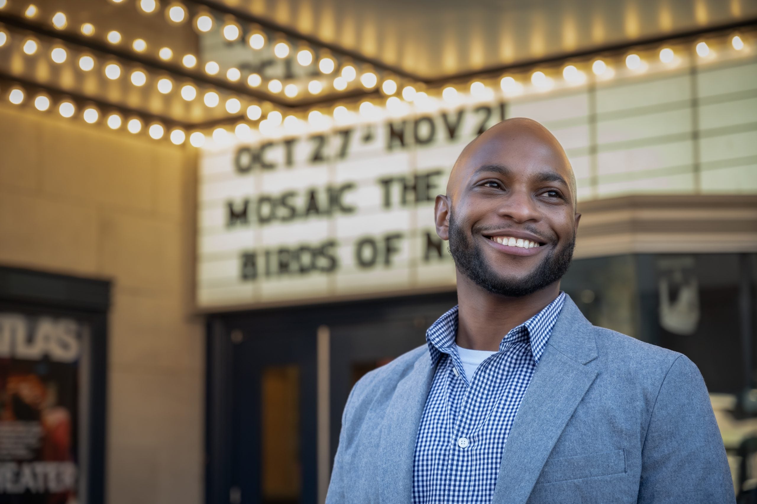 A Black male wearing a button-up shirt smiles with his hands clasped while directing a project.