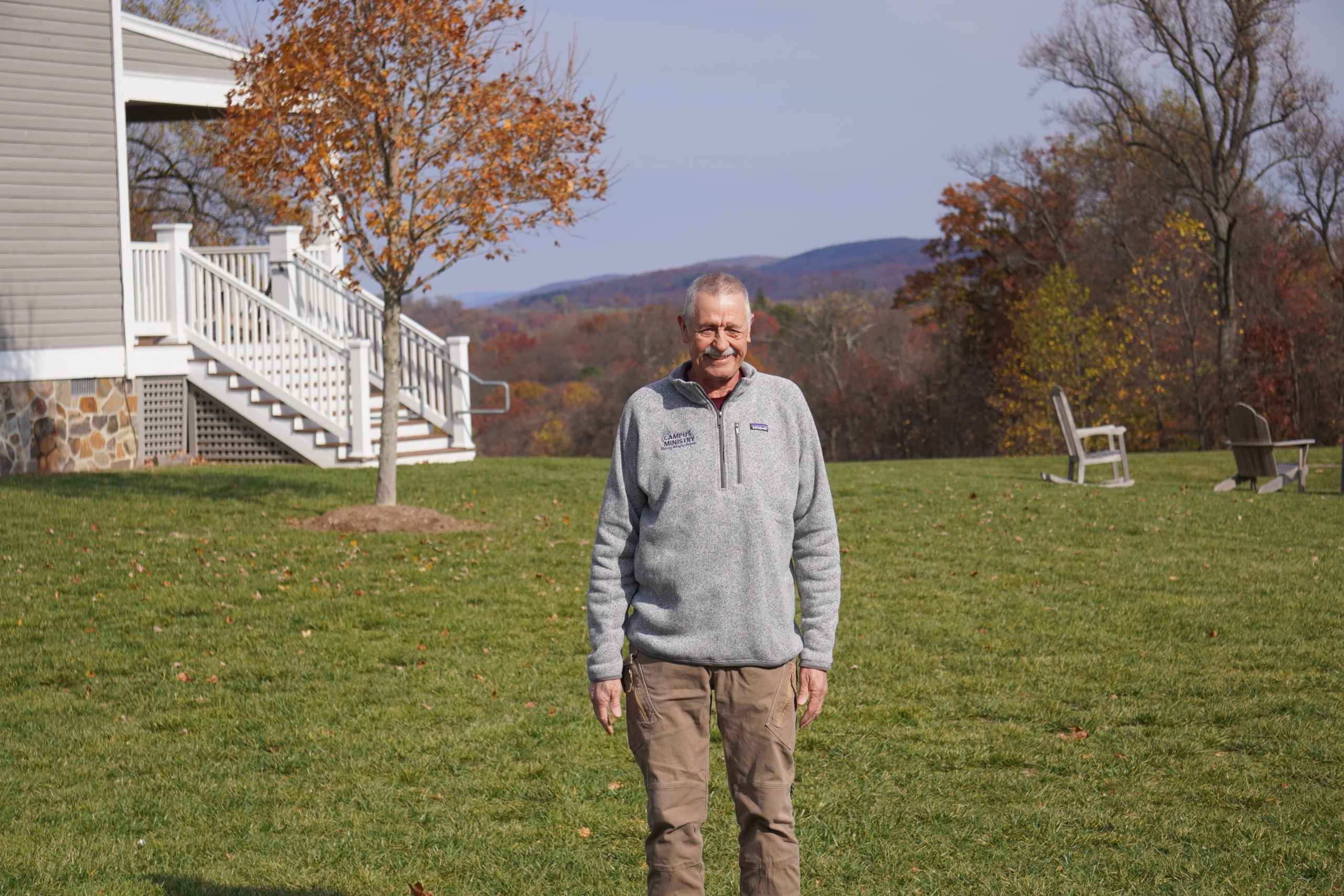 A white man wearing a gray zip-up sweater stands in front of green grass and a mountainous background.