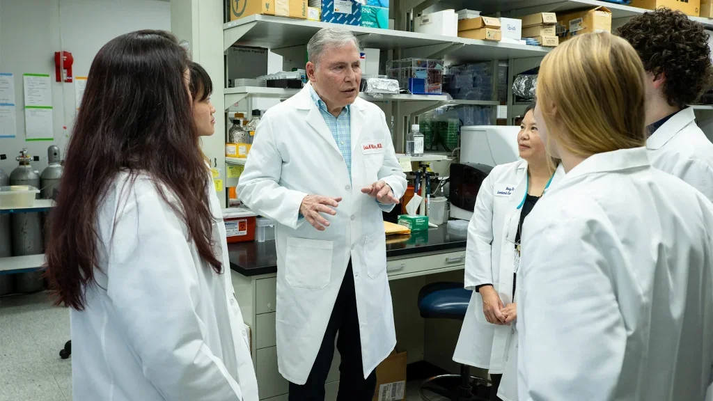 A white scientists in a lab coat stands in a laboratory talking with members of his lab, who are also wearing white coats.