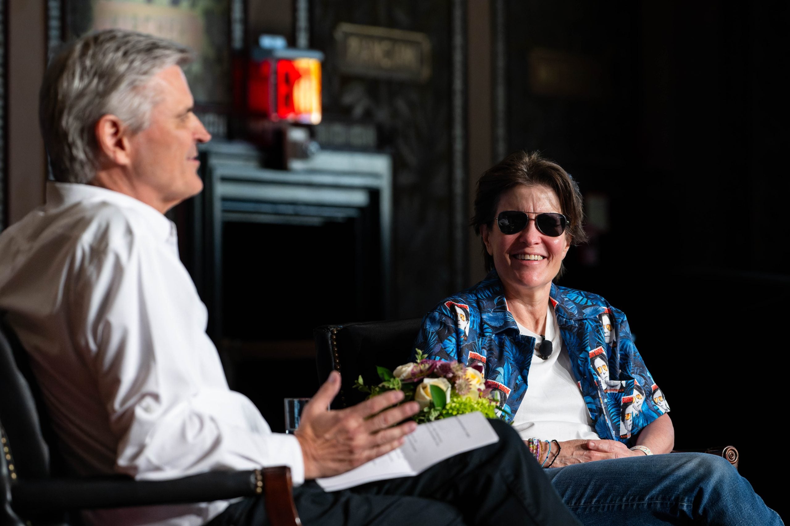 Kara Swisher, a tech podcast host and journalist, smiles on stage wearing aviator sunglasses and a blue shirt.