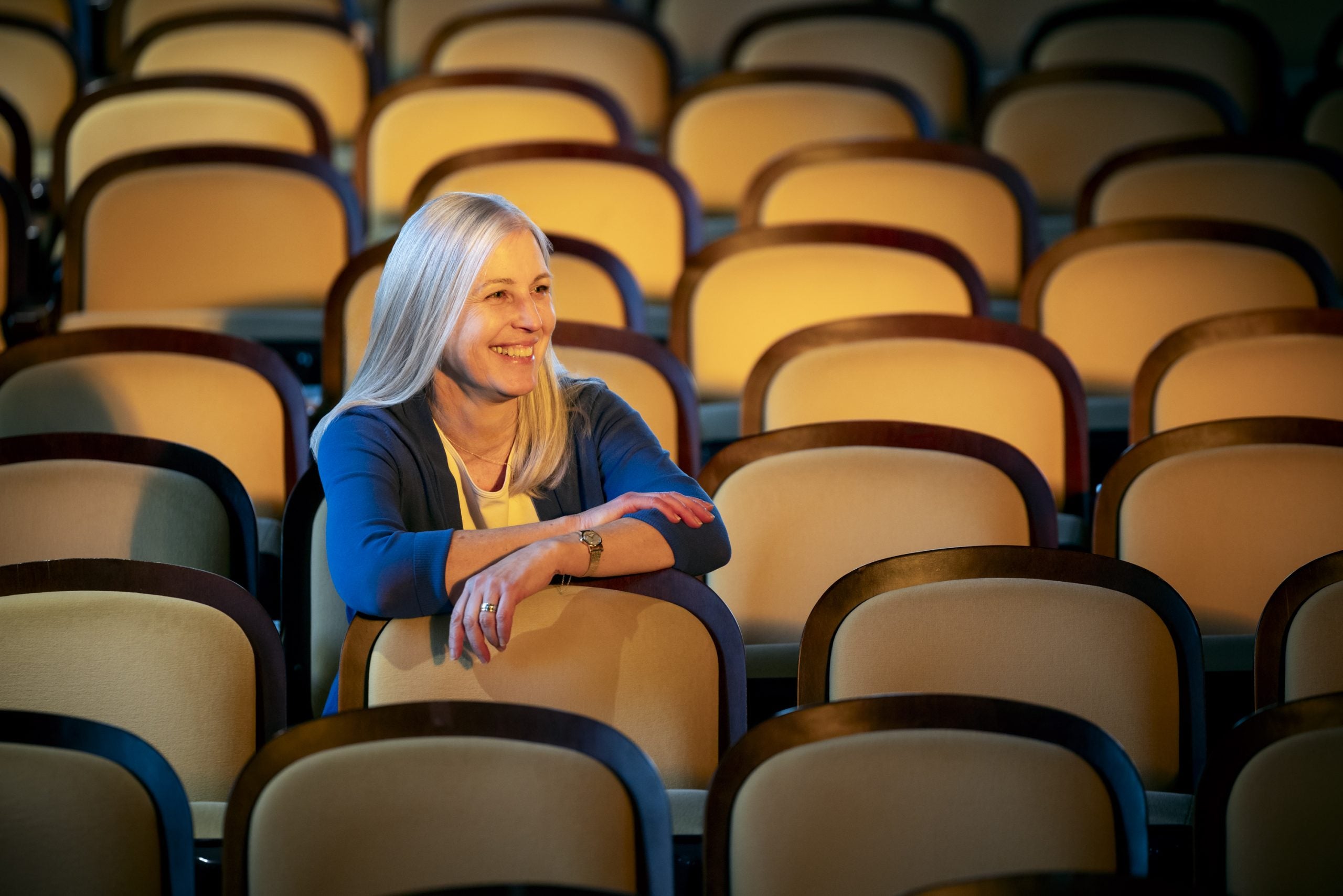 A white woman sits in an empty event space. She smiles with her arms over the seat in front of her.