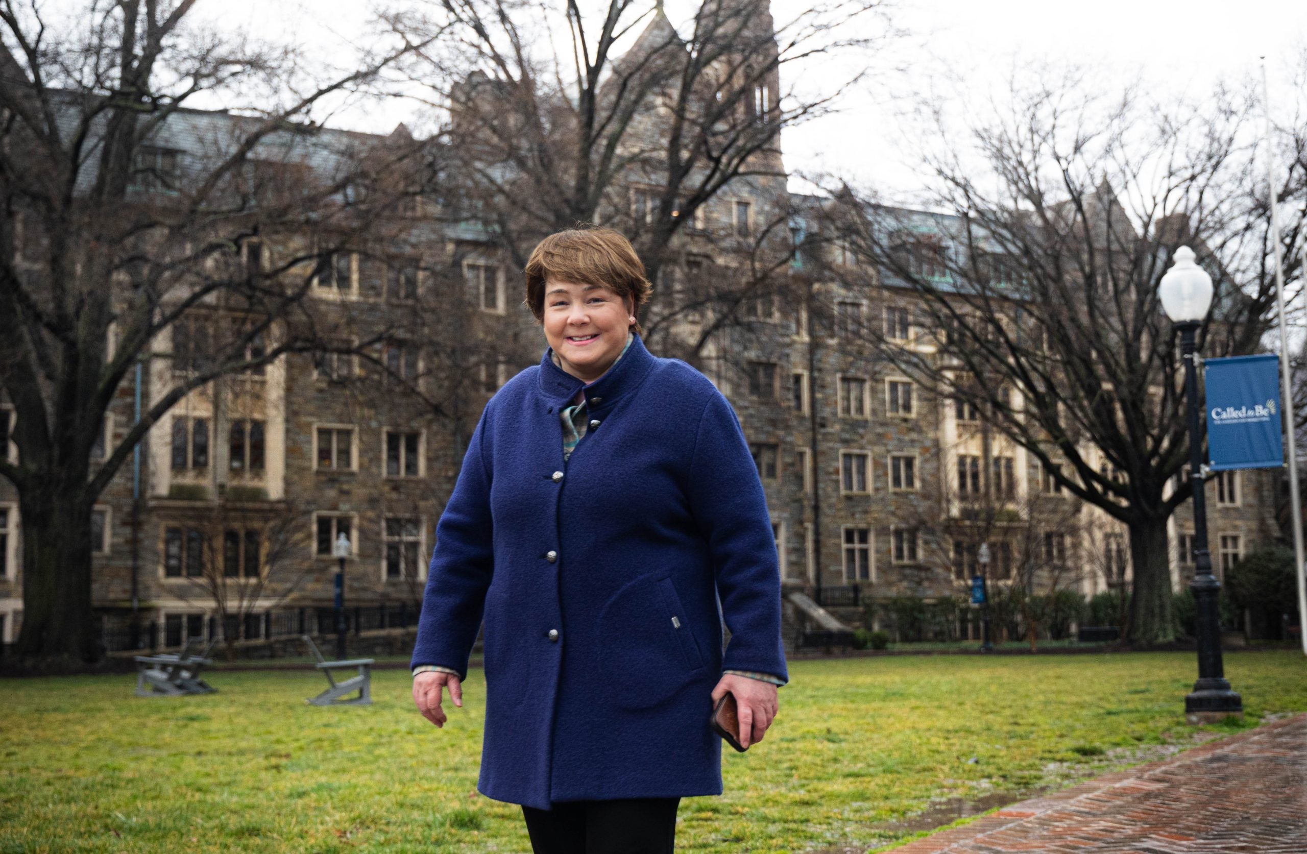 A white woman in a bright blue jacket stands in front of a stone building on Georgetown's campus smiling.