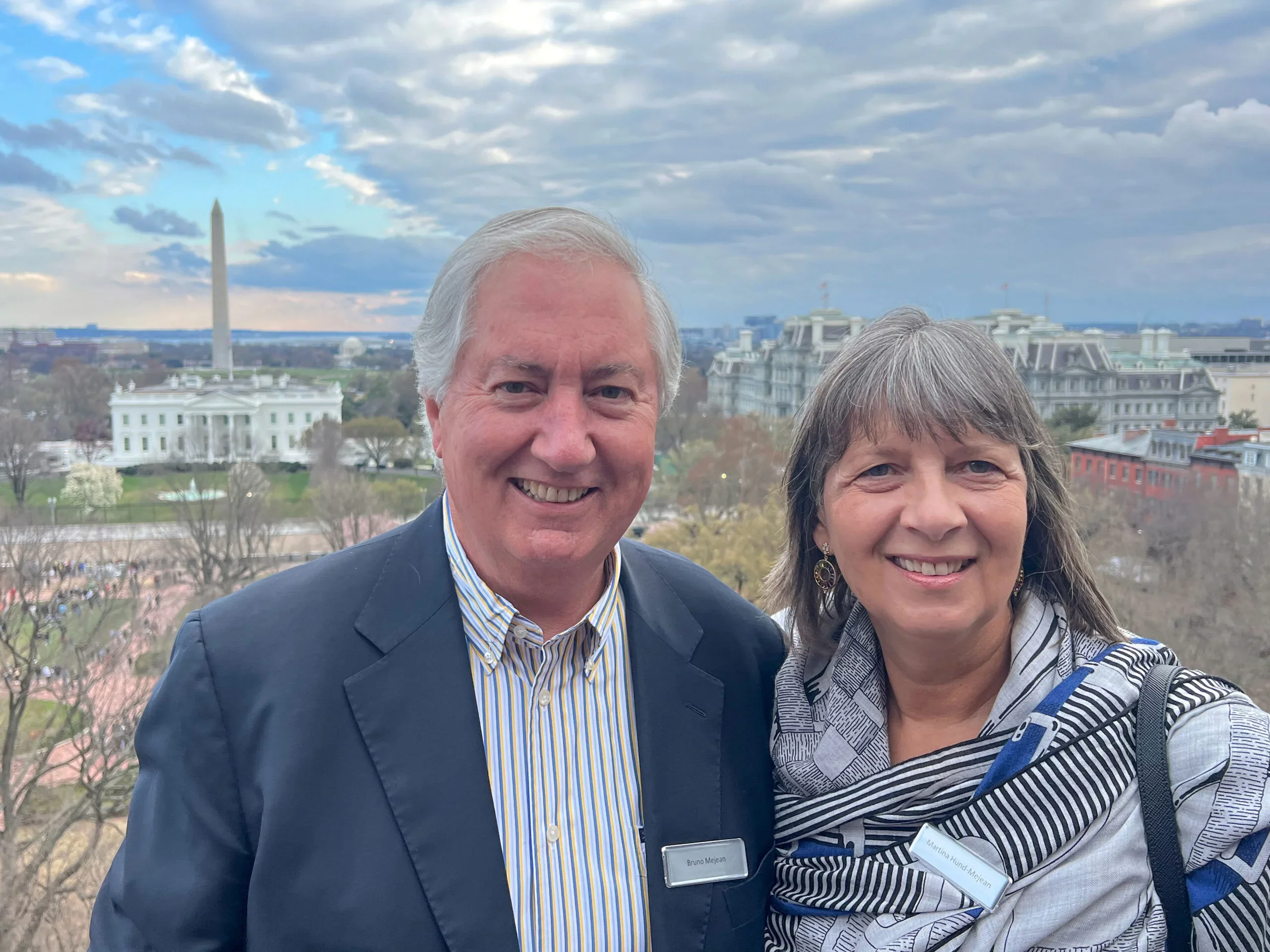 A white man and woman smile together in front of a cloudy sky and buildings.