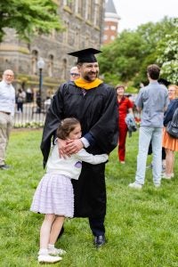A graduate in a cap and gown stands while his daughter hugs him.