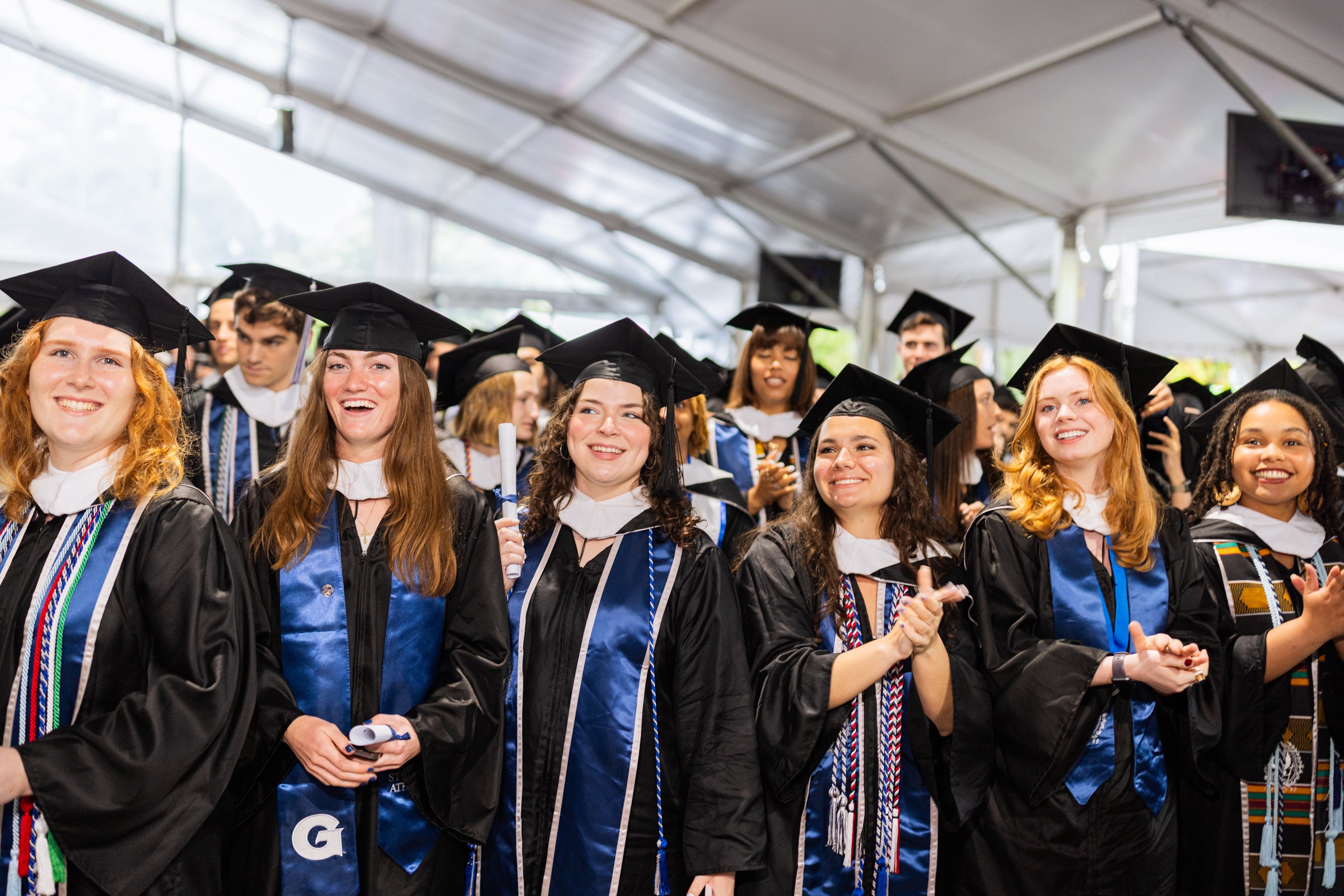 A group of graduates in caps and gowns smile at a commencement ceremony.