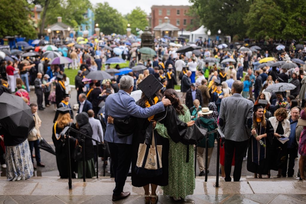A graduate and her parents stand with their arms wrapped around each other facing a crowd of commencement attendees.