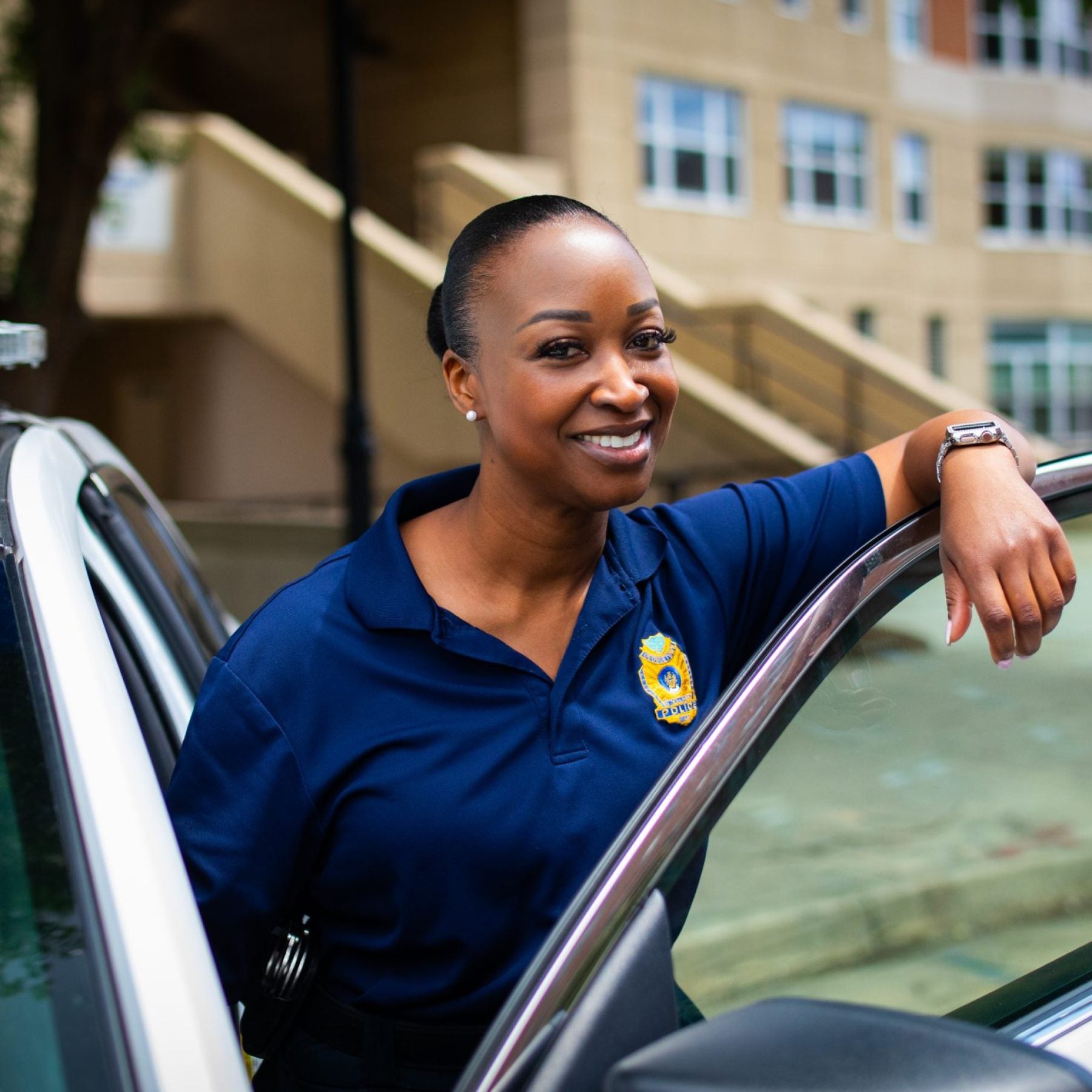 A Black female police officer leans over the door of her police car and smiles.