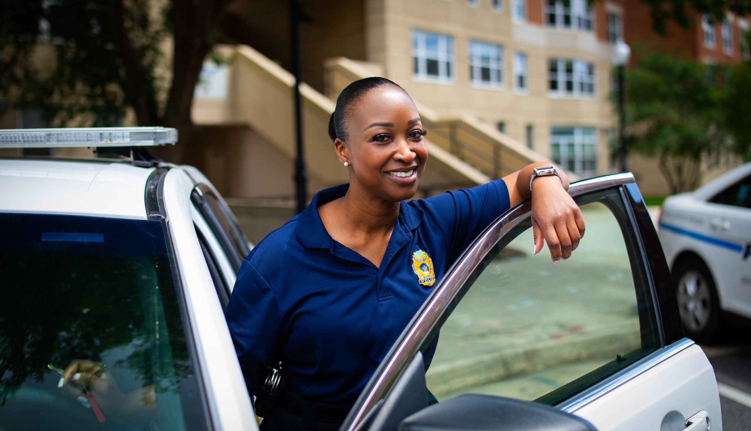 A Black female police officer leans over the door of her police car and smiles.