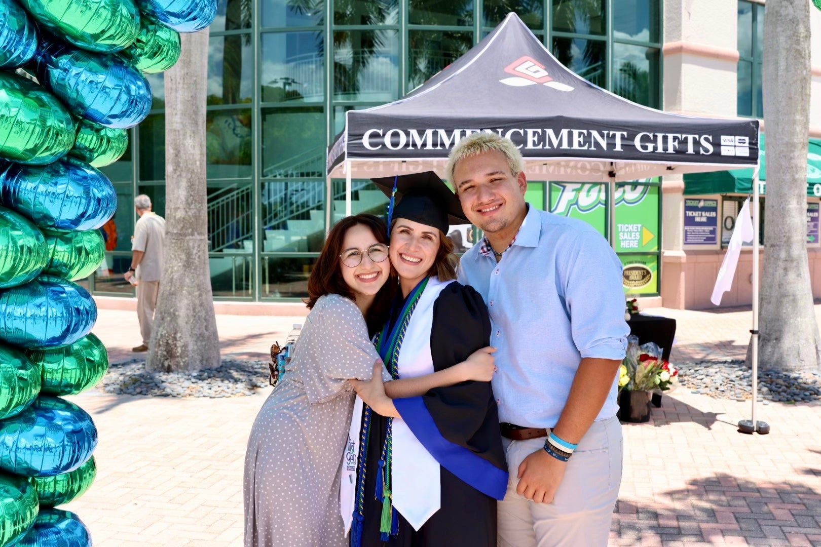 A mother graduates from college and hugs her children in front of balloons. She's wearing a cap and gown.