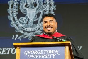 A Georgetown alumnus smiles from behind a podium wearing a graduation gown.