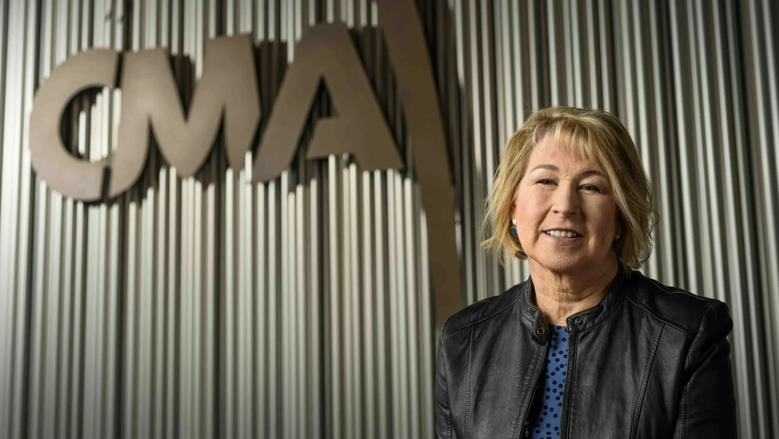 A white woman with blonde hair and a leather jacket smiles in front of a sign that says "CMA."