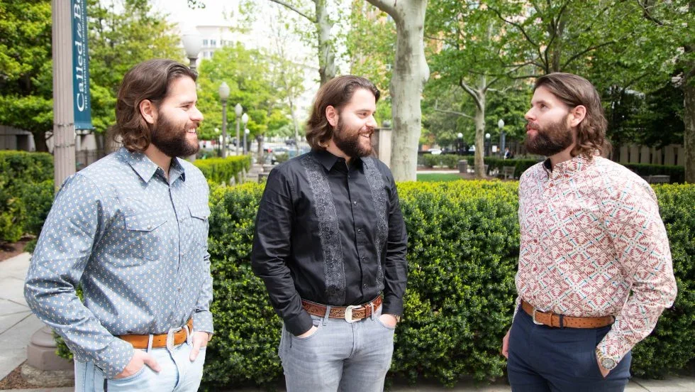 Identical triplets with brown hair and brown beards wear matching "Georgetown Law" T-shirts.